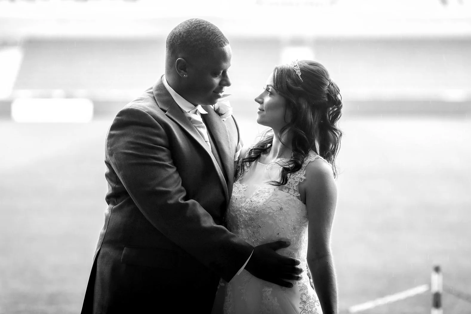 A couple in wedding attire gazing into each other's eyes on what appears to be a sports field or track, black and white photo.