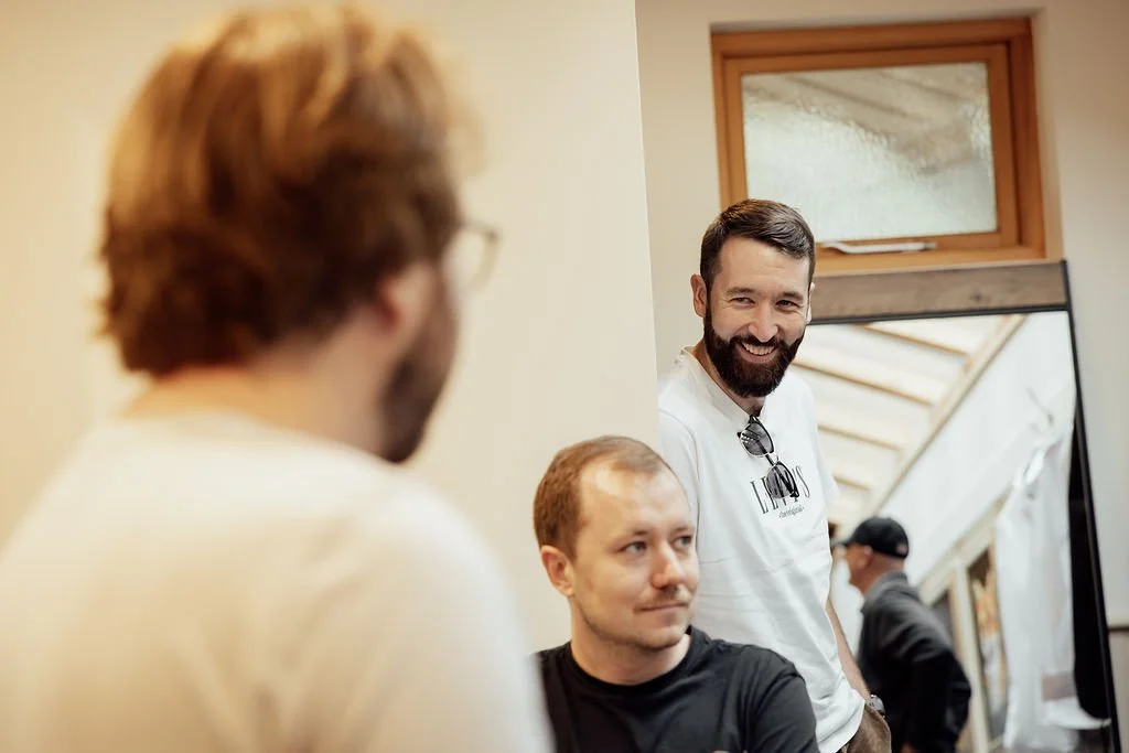 Three men having a conversation, one with glasses, the second with short hair and a beard, smiling, and the third with short hair, sitting in front of a mirror, in a room with a window.