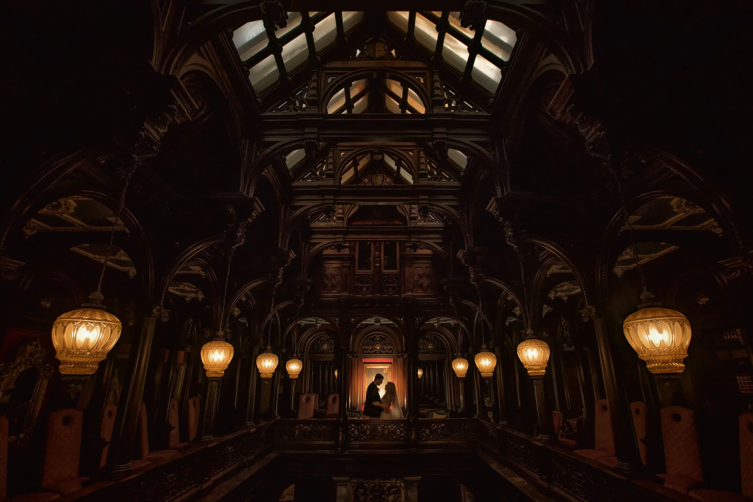 Silhouette of a couple standing close together in an ornate, darkened, gothic-style hall with intricate woodwork, large hanging lamps, and a high, glass-paneled ceiling.