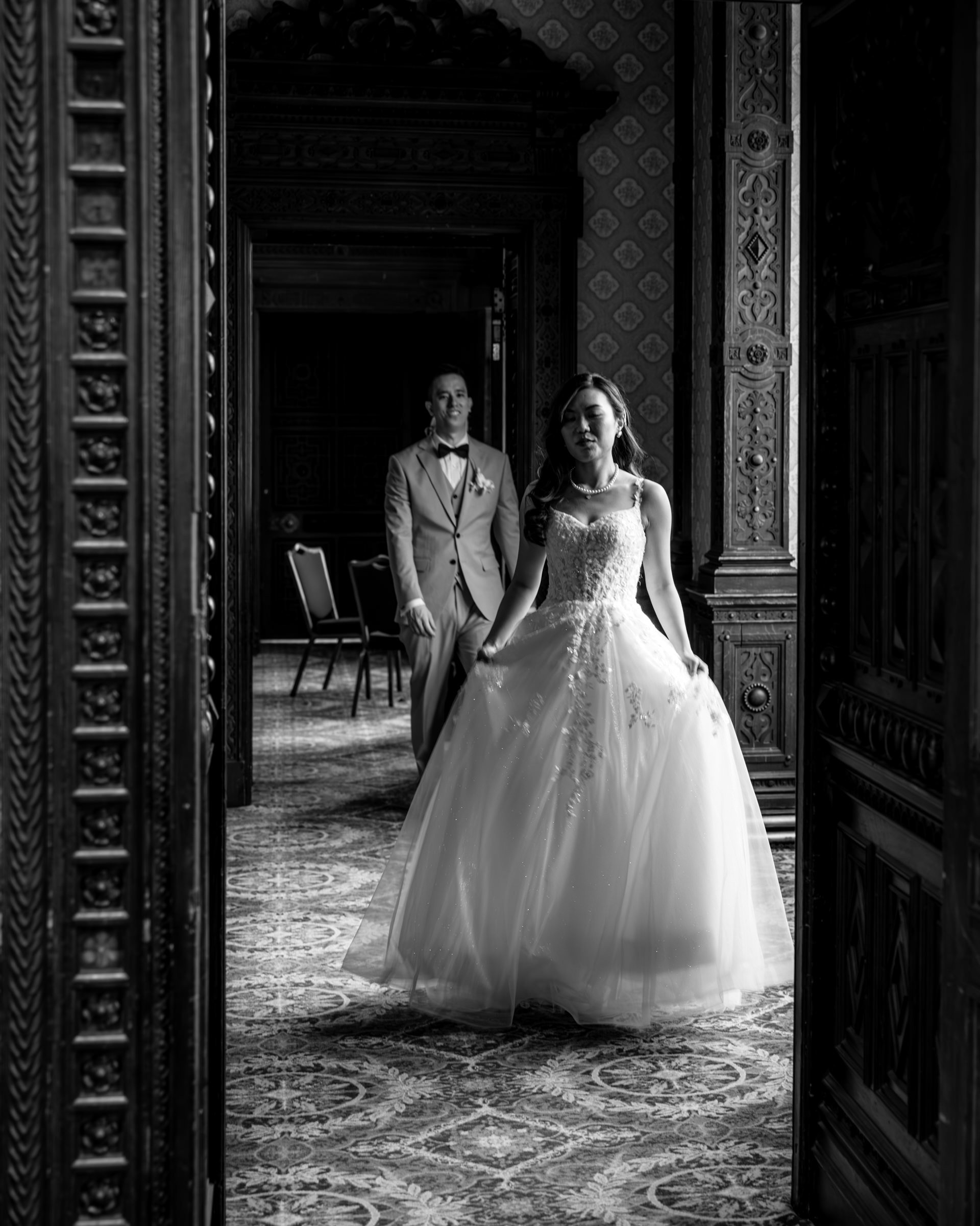 A bride in a wedding dress and a groom in a suit are standing in a grand room with ornate woodwork and patterned wallpaper.