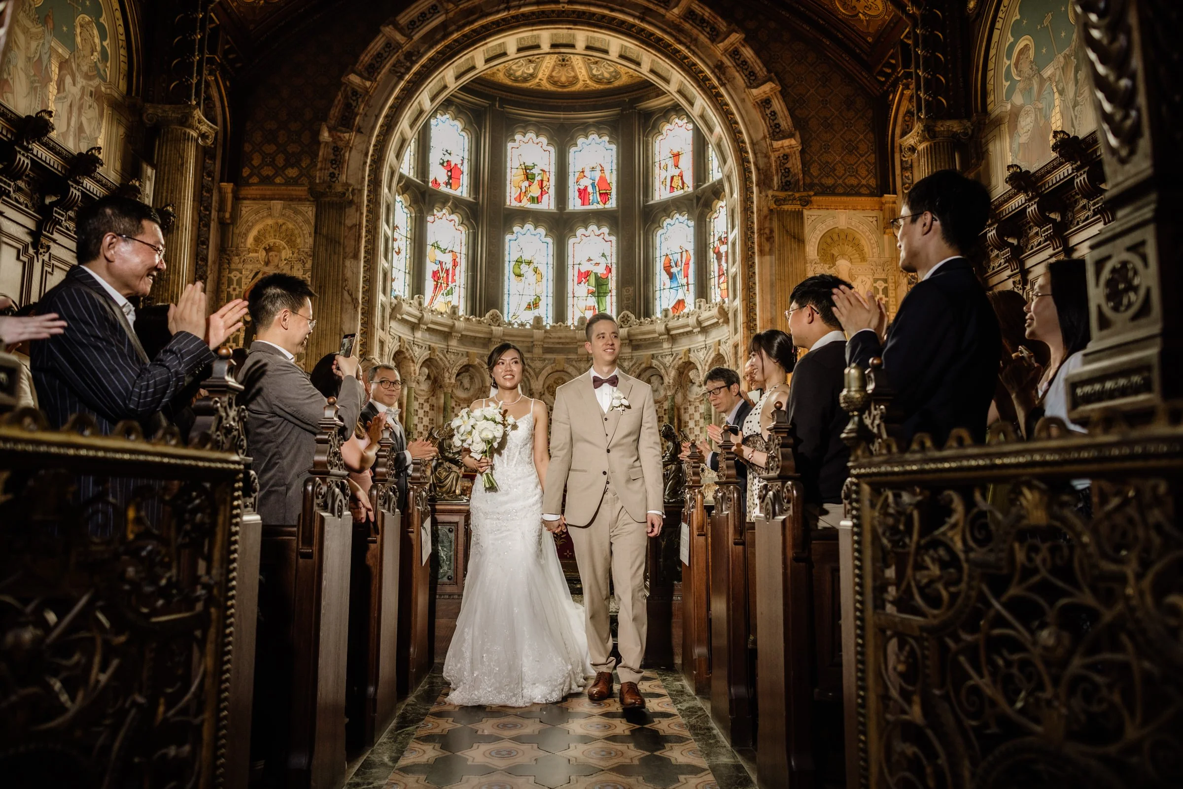 A wedding ceremony inside a church with stained glass windows. The bride and groom are walking down the aisle, holding hands. The bride is in a white gown holding a bouquet of white flowers, and the groom is in a light-colored suit with a bow tie. Guests are standing and clapping around them, some are taking photos. The church has ornate decorations and large stained glass windows depicting religious figures.