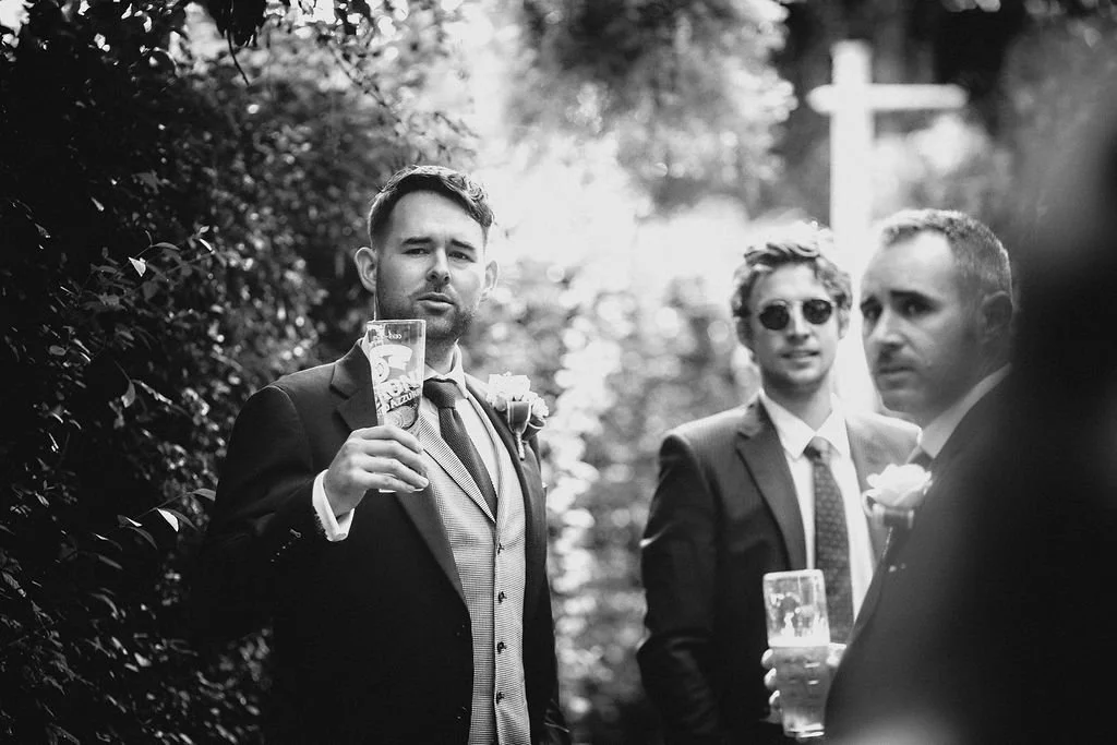 Black and white photograph of three men in suits holding glasses of beer at an outdoor event, possibly a wedding, with trees and sunlight in the background.