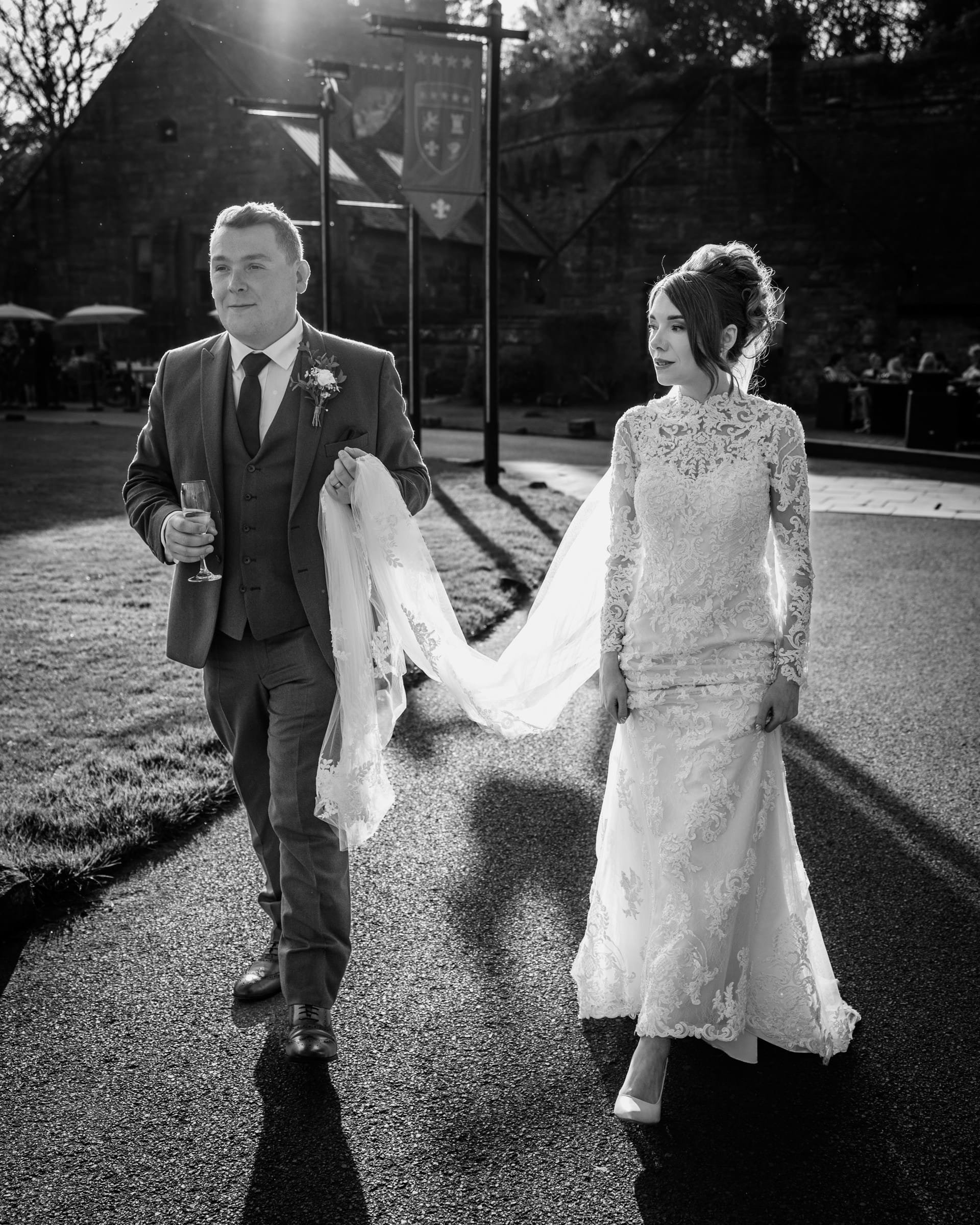 A black and white photograph of a bride and groom walking outdoors during a wedding. The groom is holding a glass of champagne and a piece of wedding fabric, and the bride is wearing a lace wedding gown and heels.