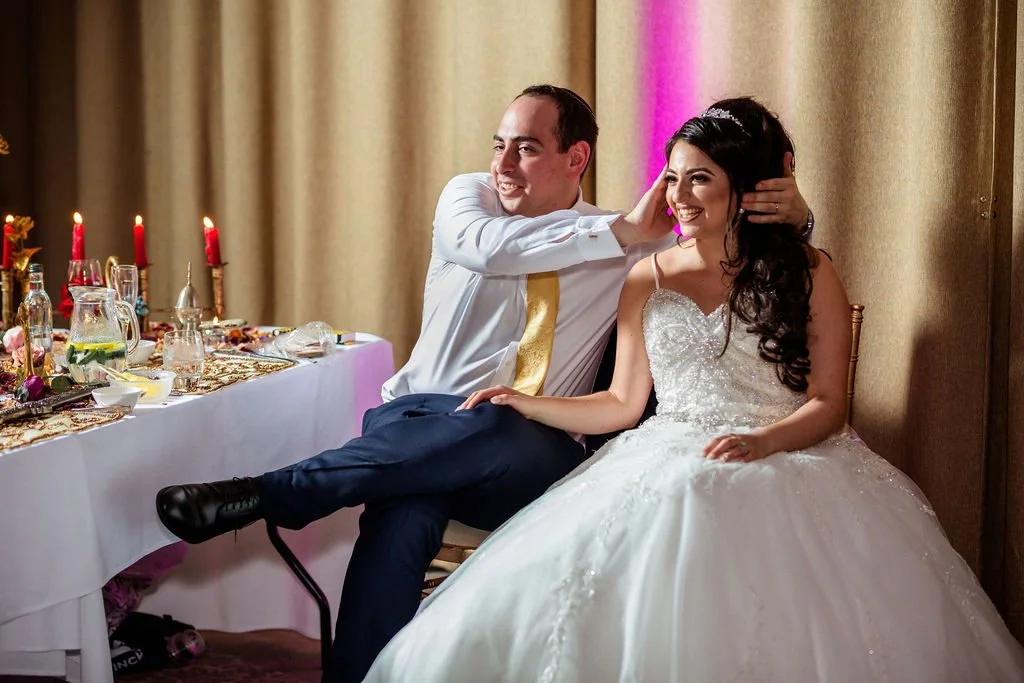 A newlywed couple sitting together at their wedding reception, the groom playfully touching the bride's head, both smiling. The bride is wearing a white wedding gown, and the groom is in a white shirt with a gold tie. There is a decorated table with 