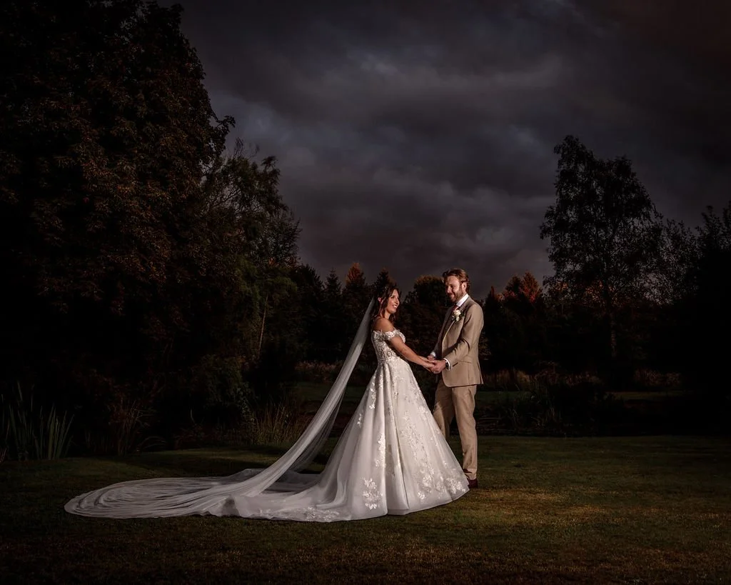 A newlywed couple holding hands outdoors during evening, with dark clouds overhead and trees in the background.