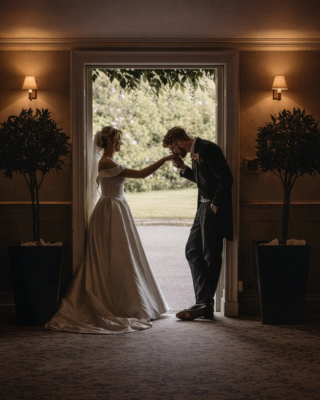 A bride and groom in wedding attire sharing a romantic moment in a doorway, with the groom kissing the bride's hand, inside a warmly lit room.