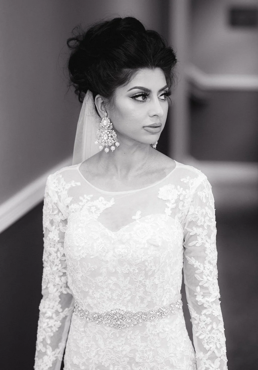 A woman in a wedding dress with lace details and large earrings, looking to the side in a black and white photo.