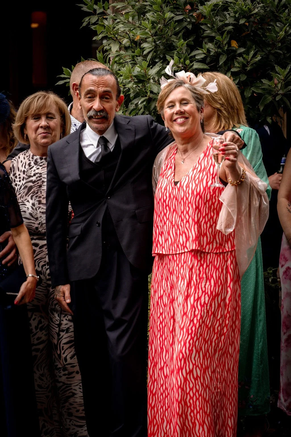 A group of people at a celebration, with a woman in a red patterned dress and a woman in a black suit holding a glass, smiling in front of greenery.
