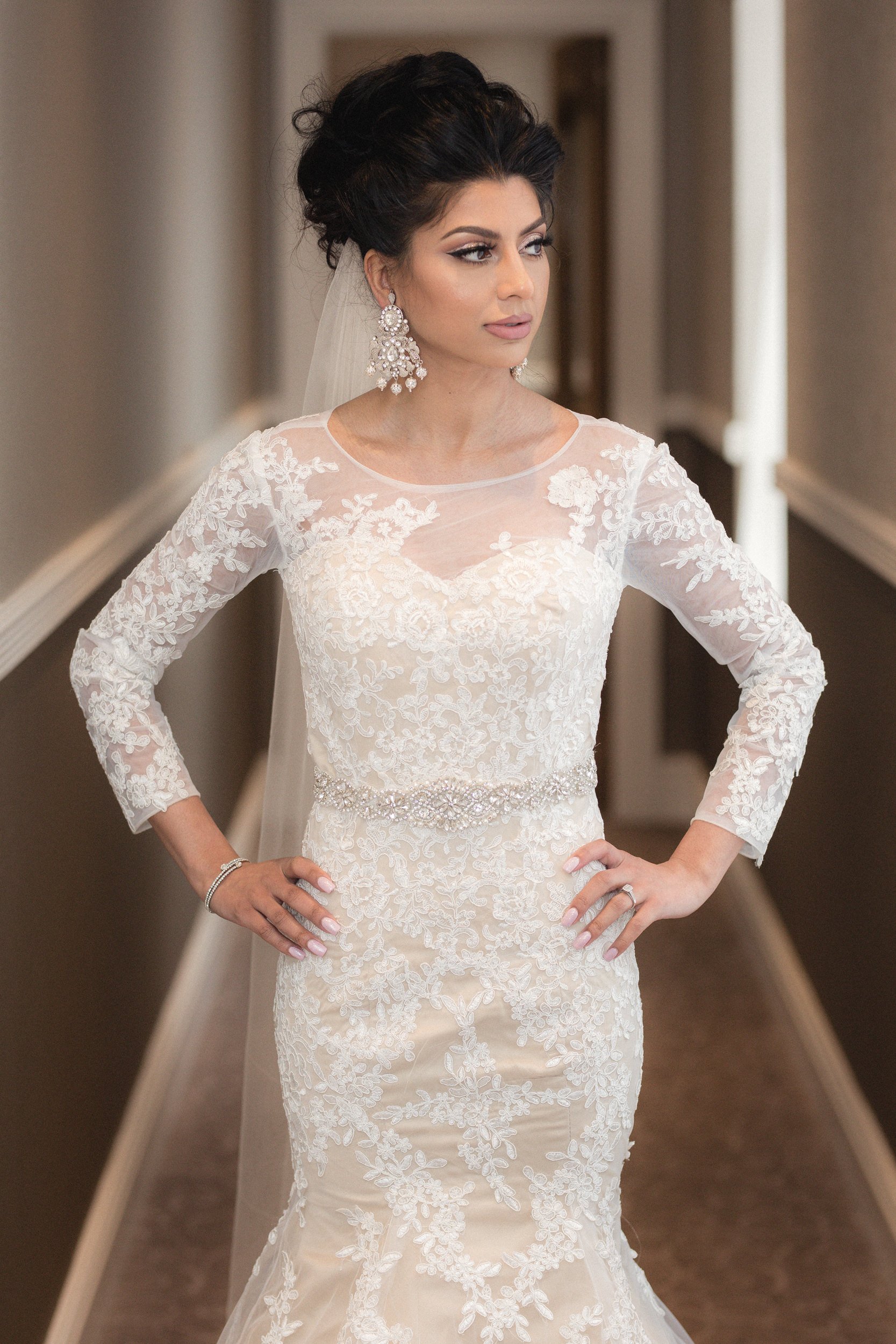 Brunette woman in a long-sleeved lace wedding gown with pearl belt, statement earrings, and a veil, standing in a hallway with her hands on her hips.
