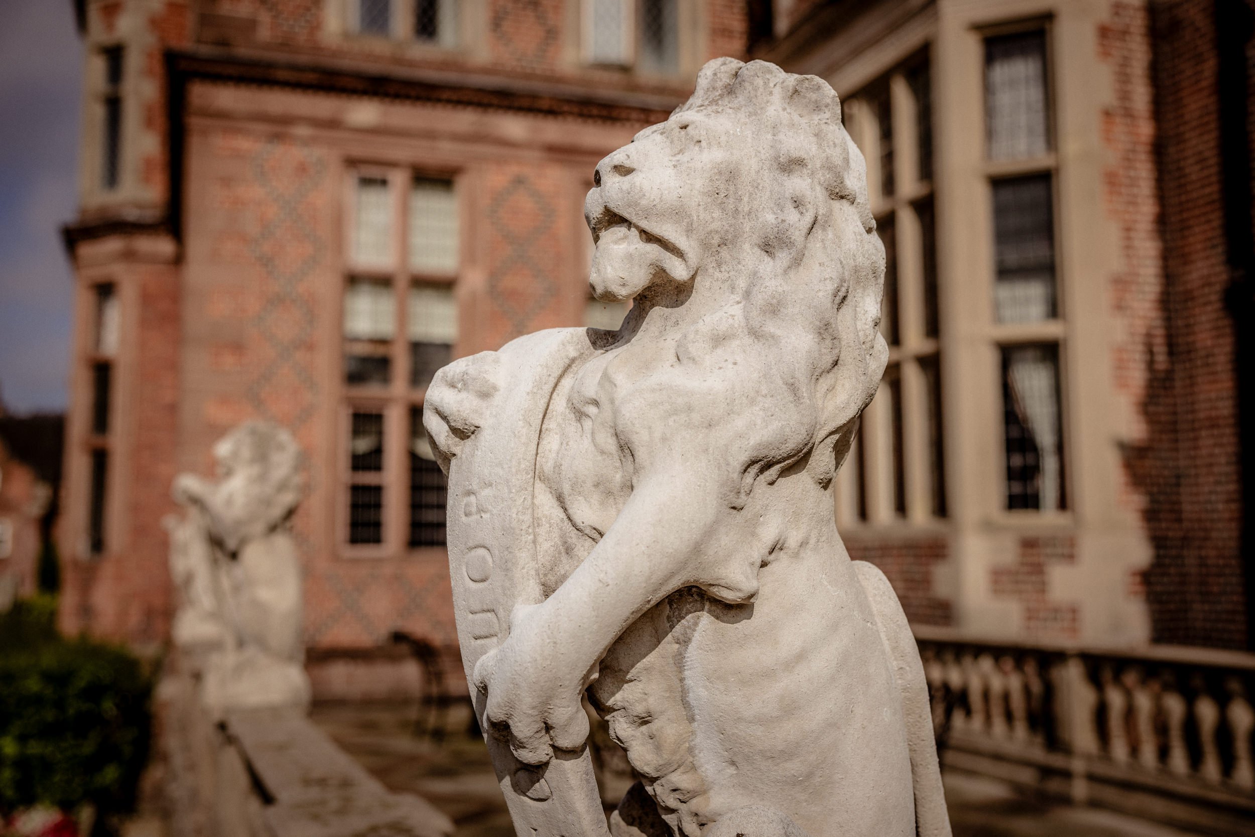 A carved stone lion statue on a pedestal outside a brick building with decorated windows. Another lion statue is visible in the background.