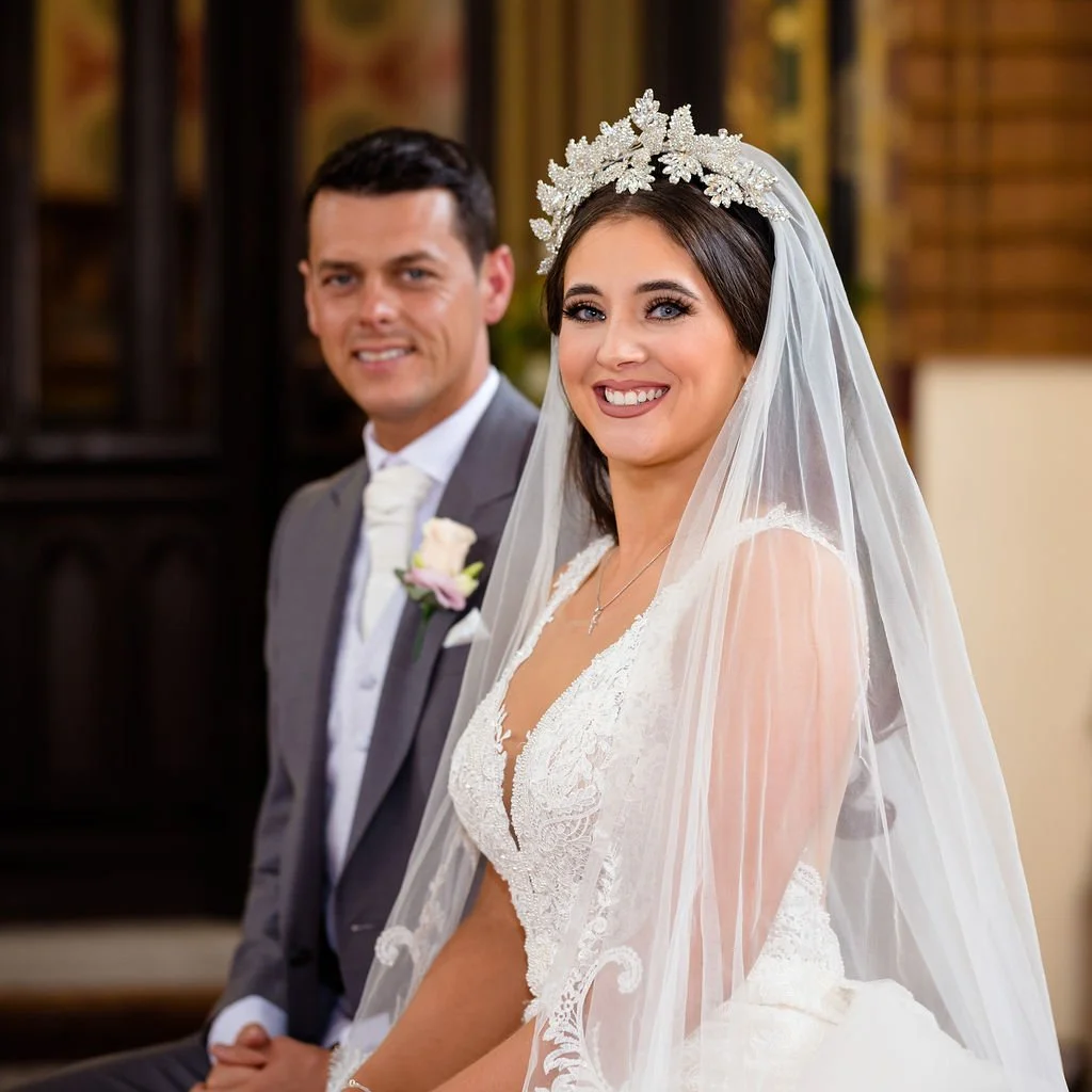 A bride and groom smiling, with the bride wearing a white lace wedding dress, veil, and floral headpiece, and the groom in a gray suit with a white shirt and tie, indoors with warm lighting.