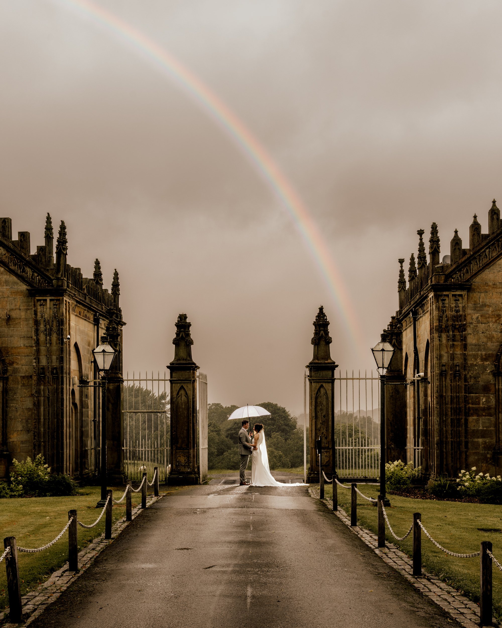A bride and groom standing under an umbrella in front of an open wrought-iron gate, with a rainbow in the sky and historic building facades on either side.