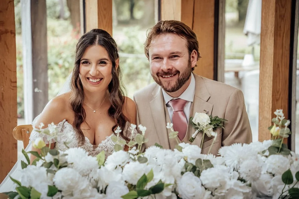 Happy bride and groom sitting at a wedding reception table decorated with white flowers inside a rustic venue with large windows.