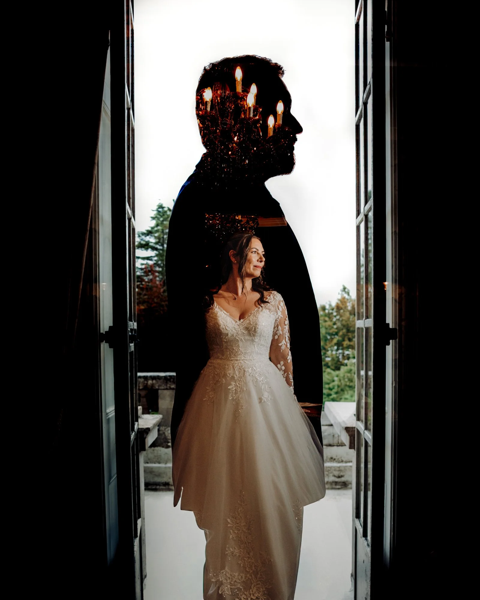 A woman in a wedding dress standing in a doorway with a silhouette of a man with candle-like lights above his head reflected in a large glass window behind her.