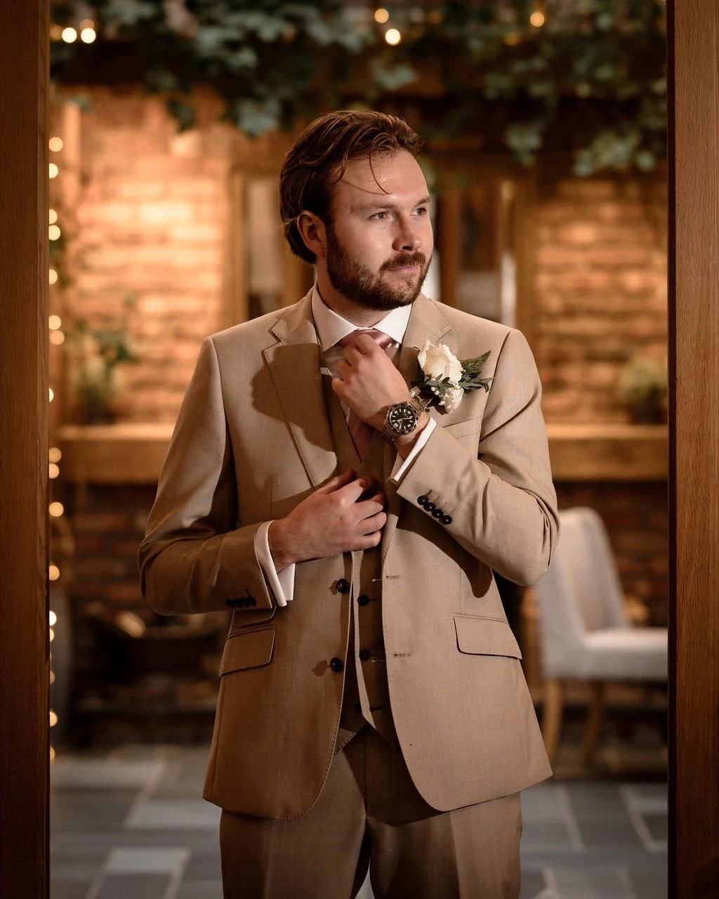 A man with brown hair and a beard stands in a warmly lit room, adjusting his beige suit jacket and wearing a white shirt, a wristwatch, and a white boutonniere. He appears thoughtful, with a background of wooden walls and decorative lighting.
