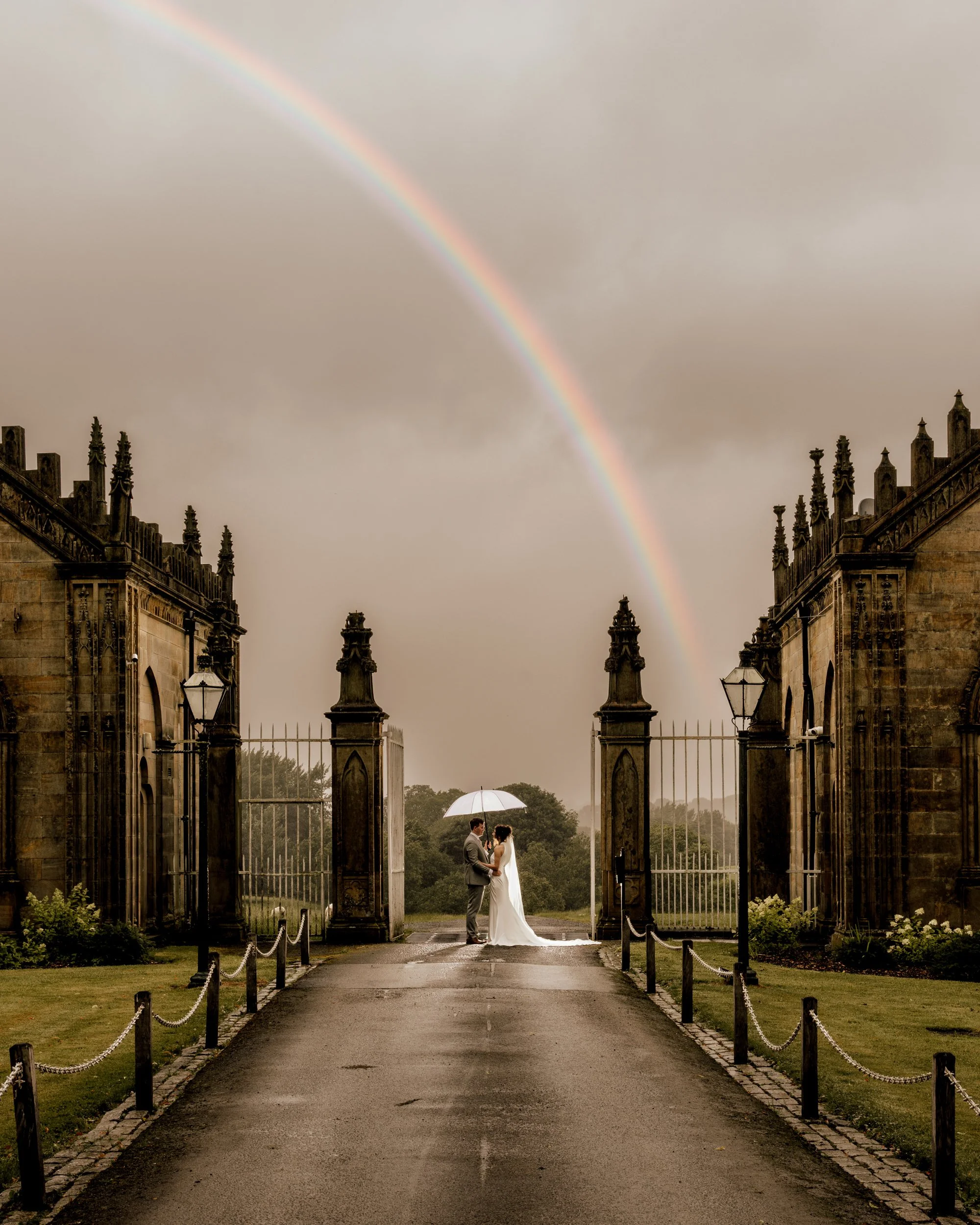 A bride and groom standing beneath an umbrella, holding hands, in front of a decorative gate with a rainbow in the cloudy sky overhead.