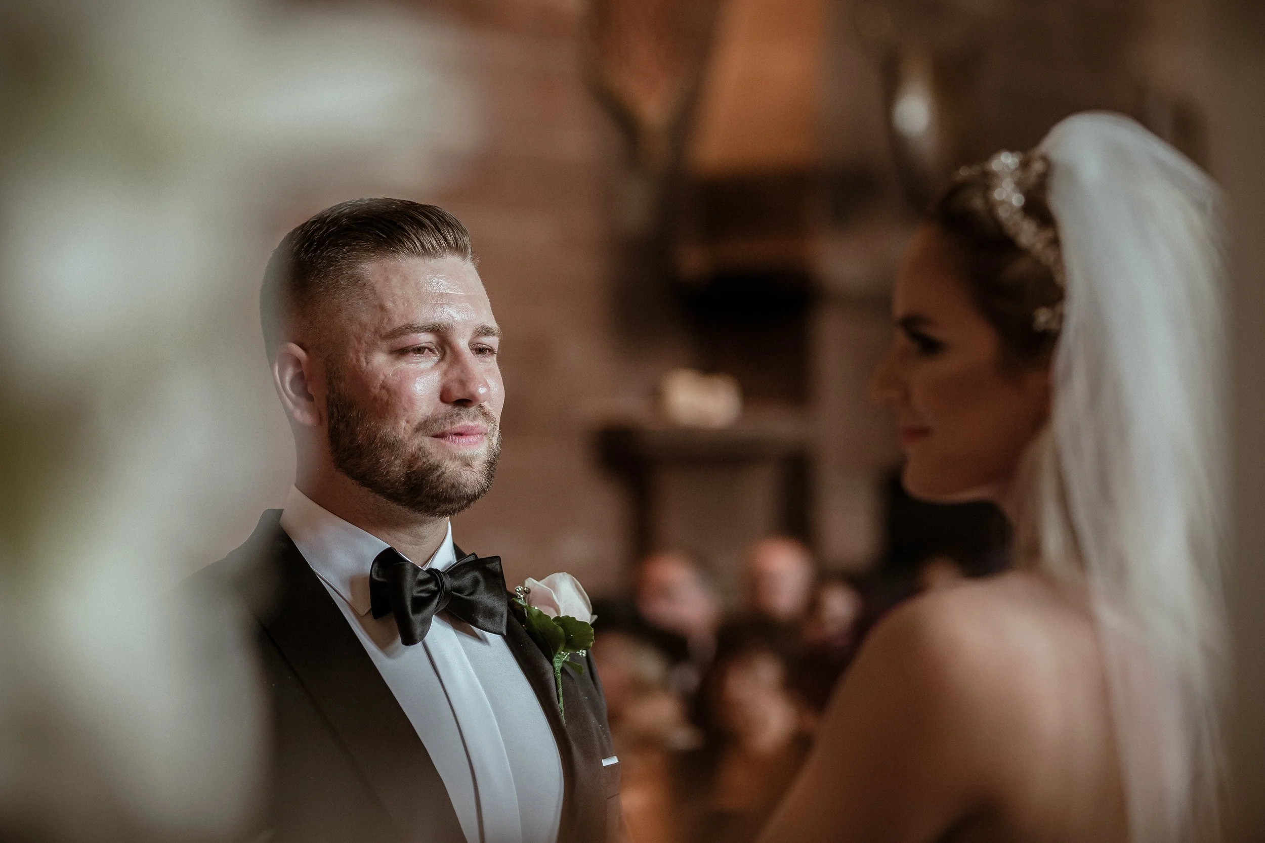 A groom and bride during their wedding ceremony, facing each other inside a warmly lit venue.
