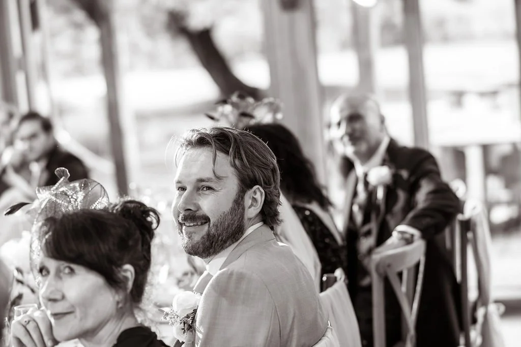 People seated at a table during a wedding celebration, smiling, in an indoor setting with large windows.