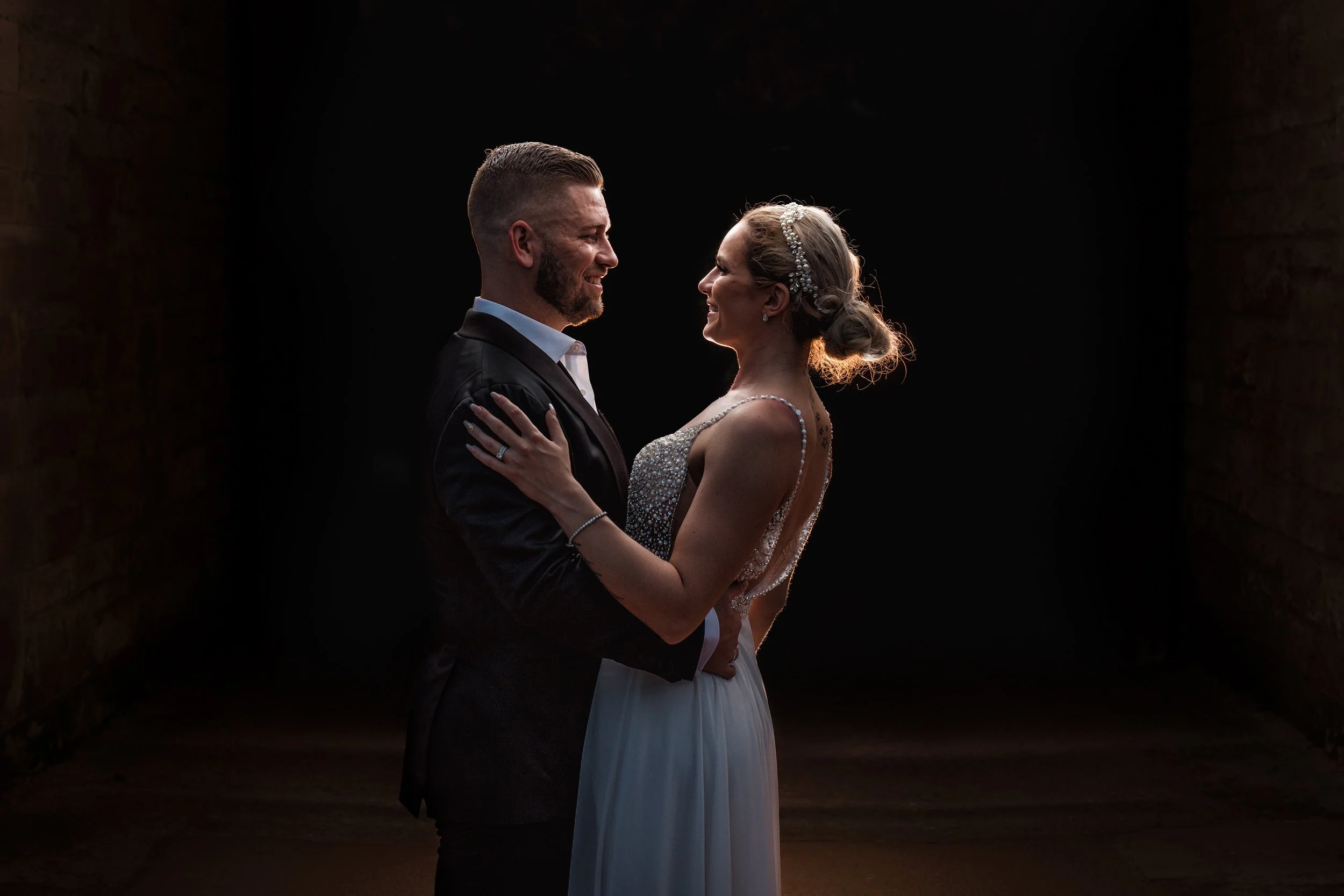 A bride and groom standing close together in a dark setting, gazing into each other's eyes, with the bride wearing a sparkly dress and the groom in a suit.