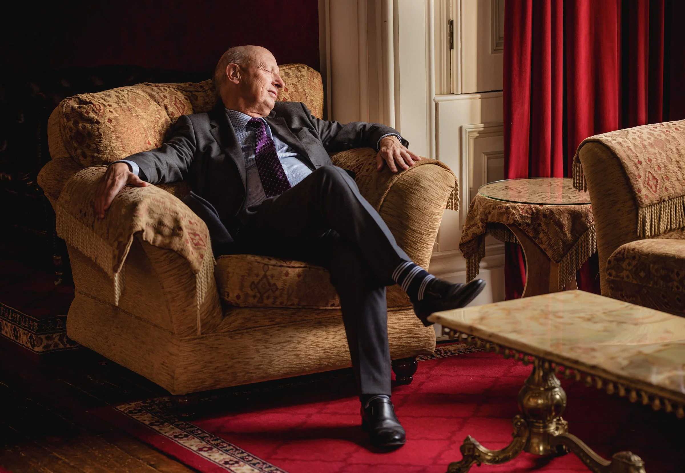 An elderly man in a suit sitting on a patterned armchair in a luxurious room, relaxing with his eyes closed, with his arms resting on the armrests and one leg crossed over the other.