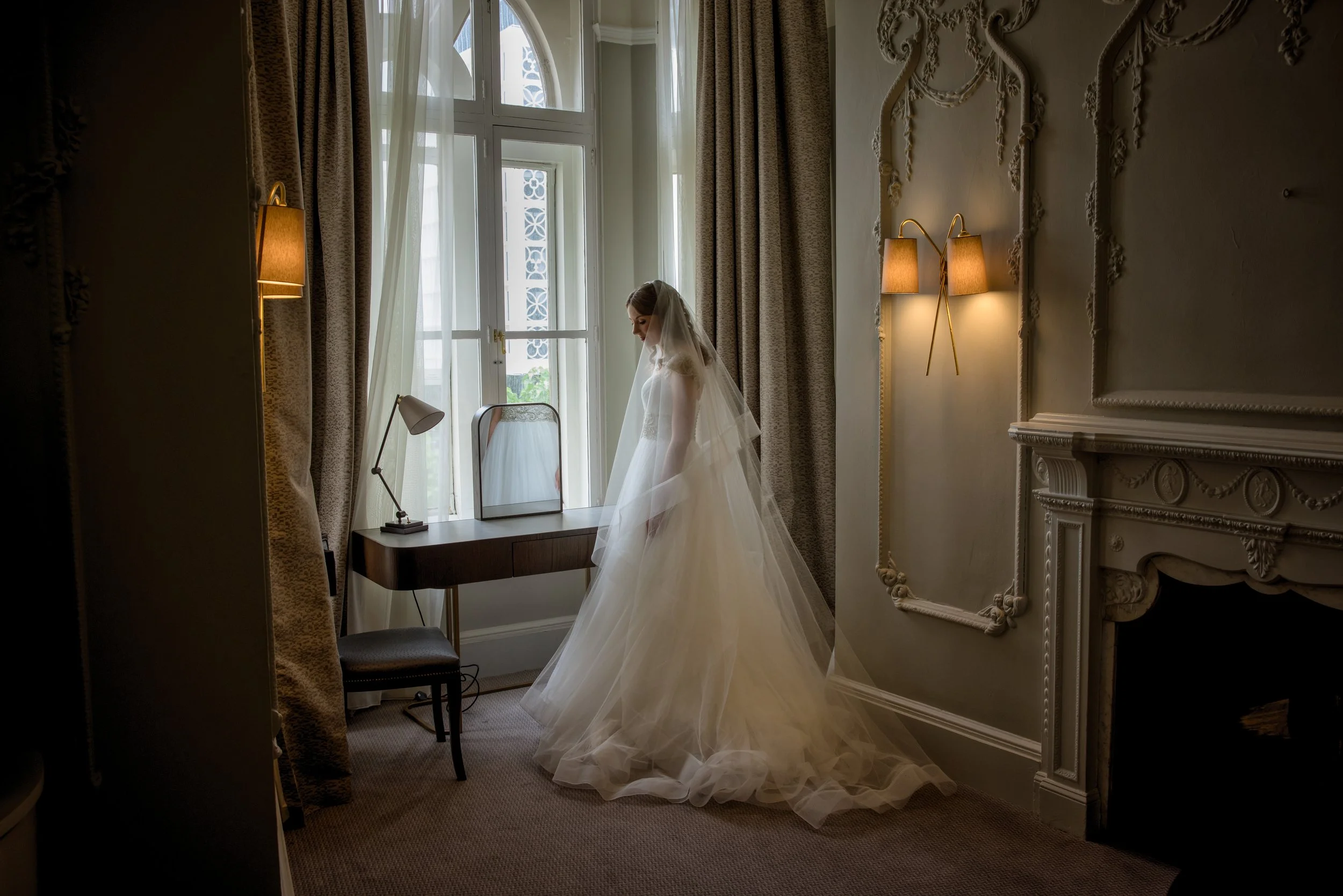 Bride in white wedding dress pensively stands by the window in her Manchester town house room.