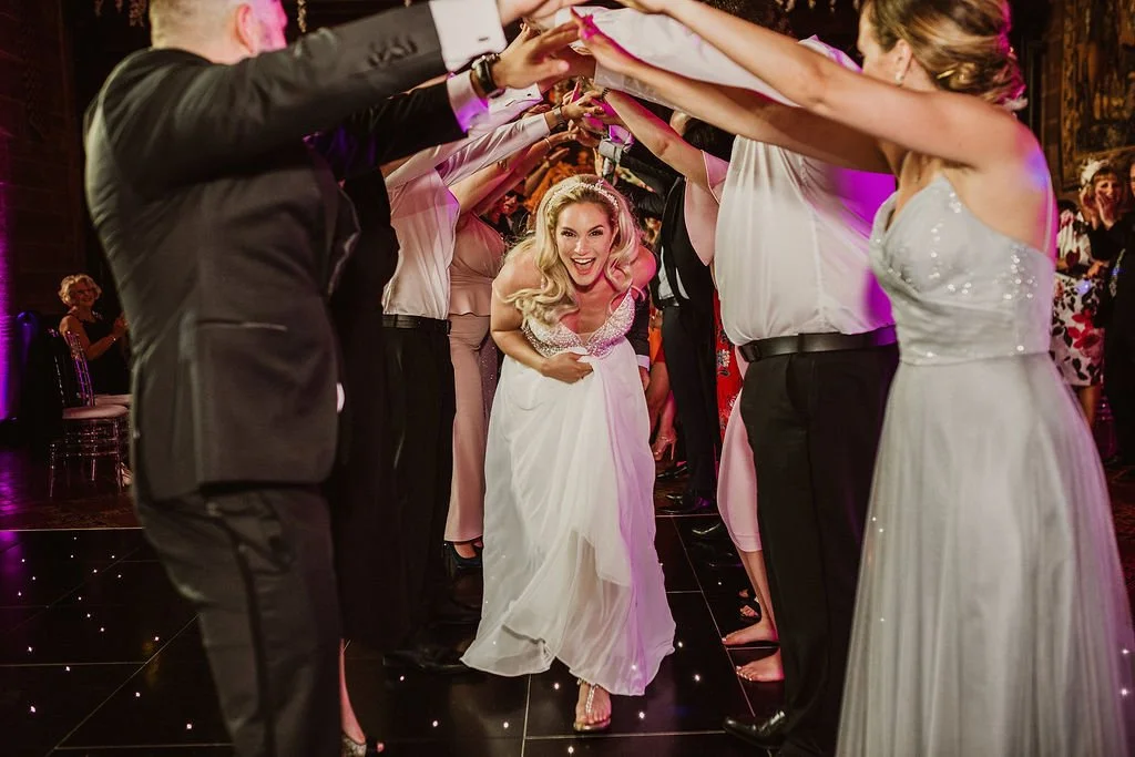 Bride in a white wedding gown smiling and walking through a tunnel of raised hands at her wedding reception.