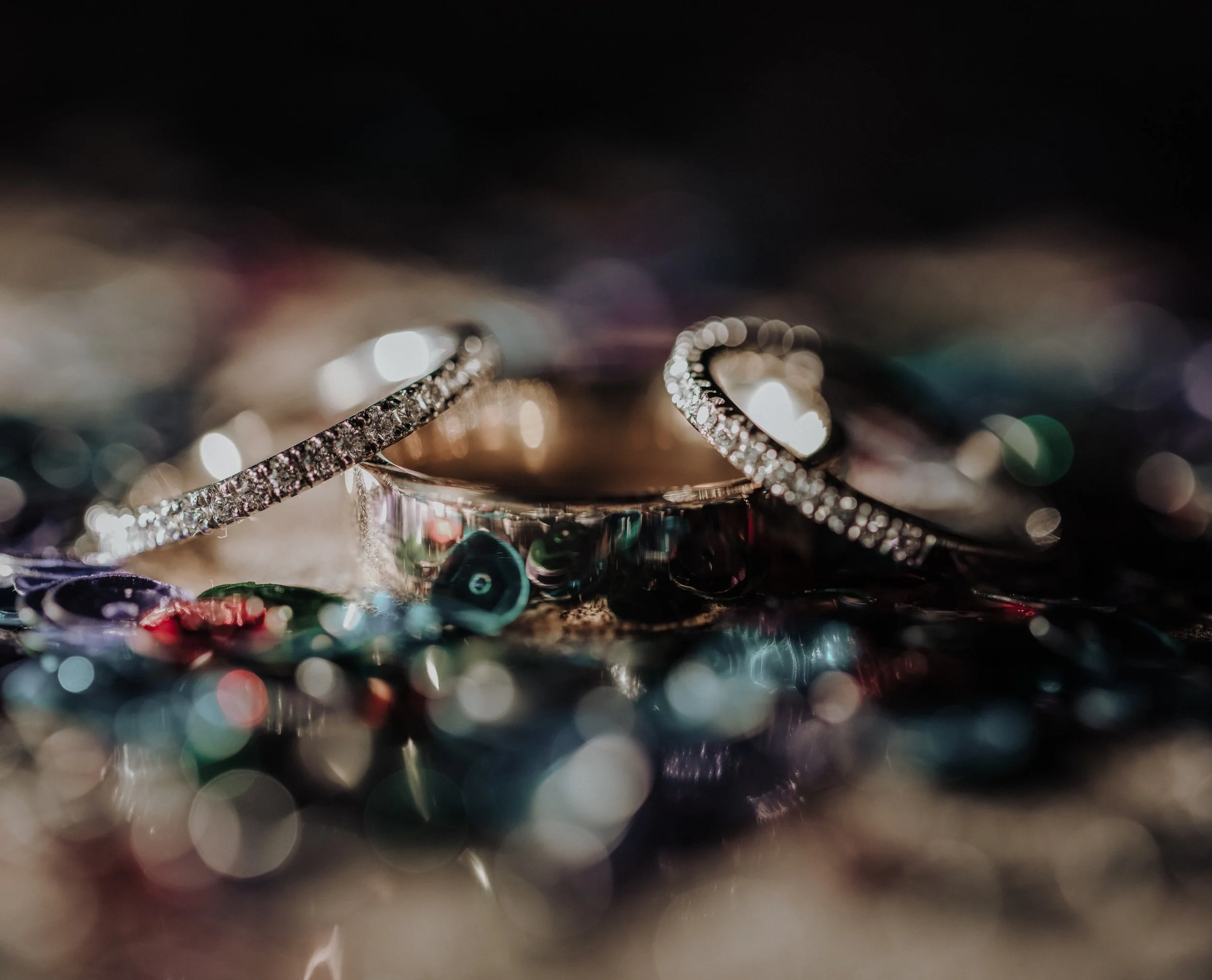 Close-up of two diamond-studded rings on a multicolored, blurred reflective surface with bokeh effect.