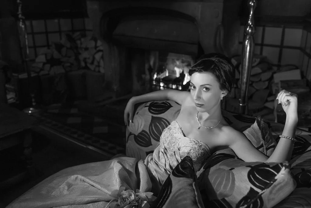 A woman with short dark hair wearing a strapless dress sitting on a patterned sofa near a fireplace, with logs stacked nearby and a lit fire inside.