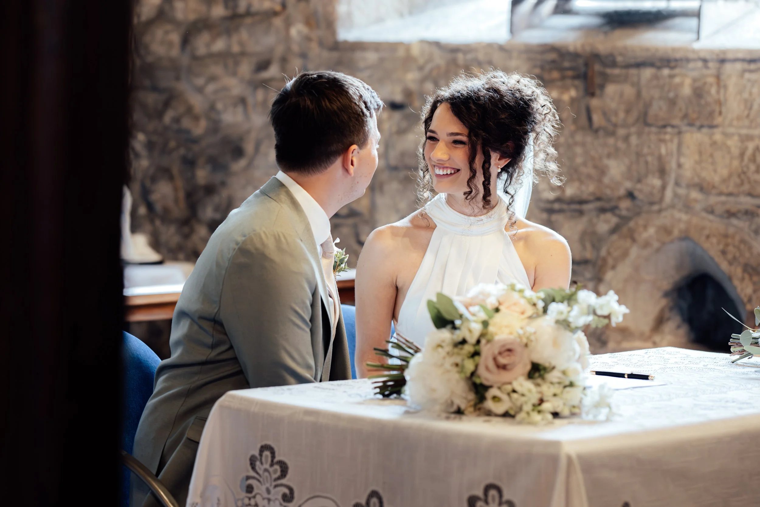 A bride and groom sit across from each other at a wedding reception table, smiling at each other, with a bouquet of flowers on the table and a brick wall in the background.