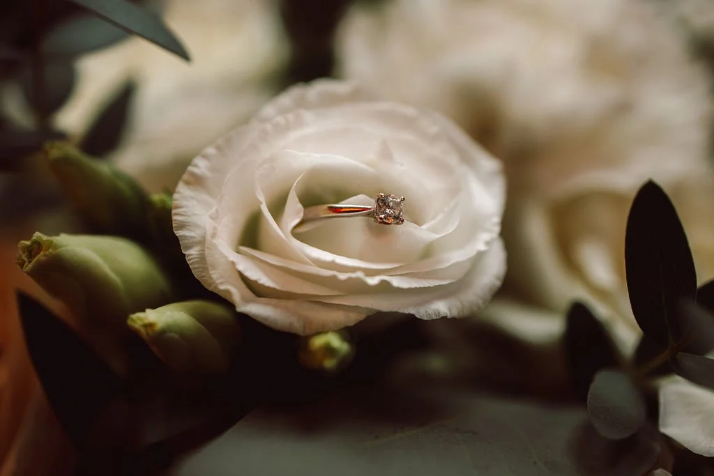 A diamond engagement ring on a white flower surrounded by green buds and dark leaves.