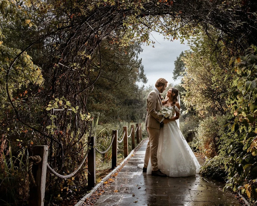 A bride and groom standing on a wooden pathway surrounded by lush greenery, embracing each other happily on their wedding day.