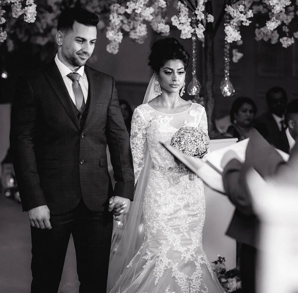 A black and white photo of a wedding ceremony featuring a bride and groom holding hands. The bride is wearing a lace wedding dress and holding a bouquet, while the groom is dressed in a suit. They are standing in front of an officiant, with guests in