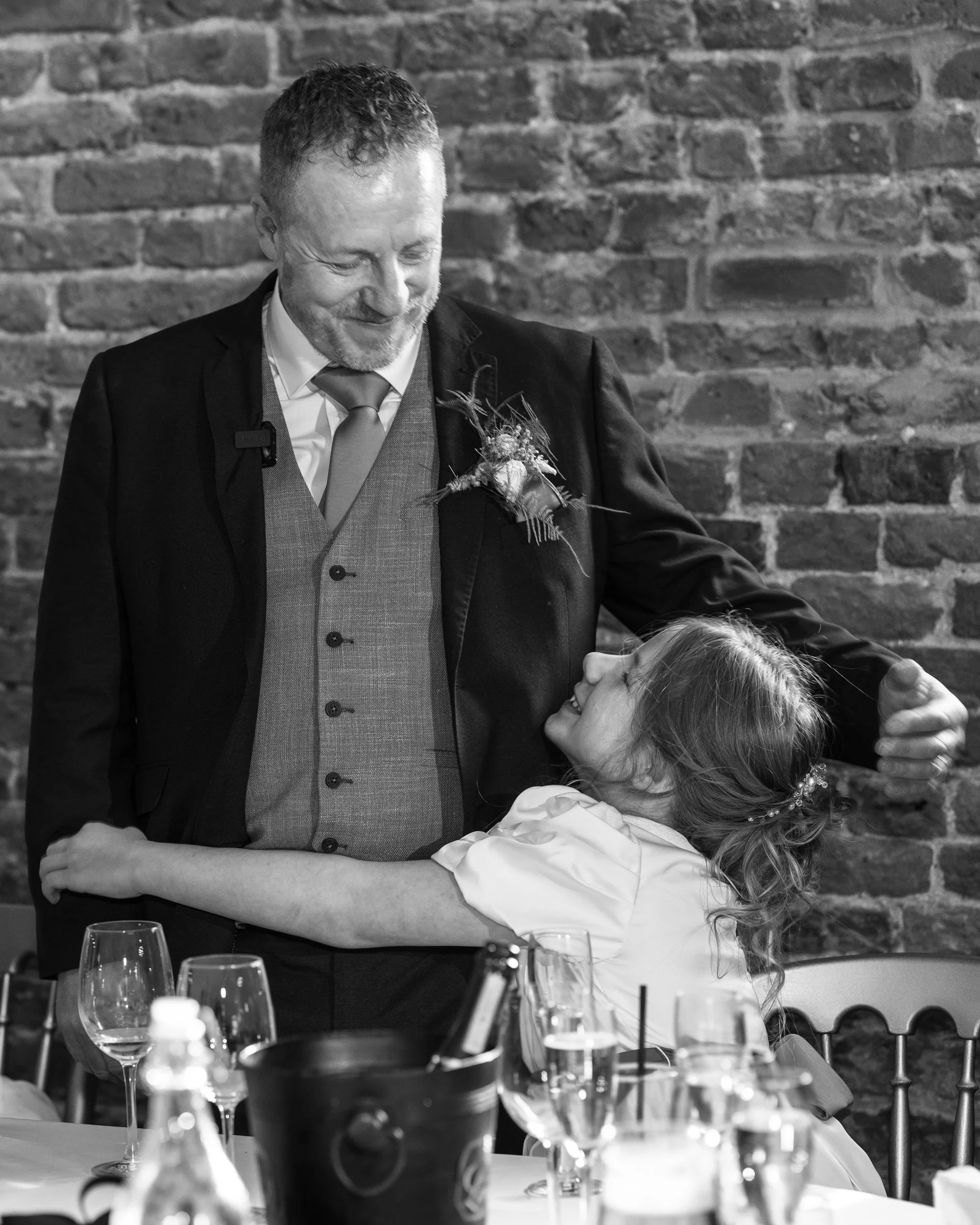 A man and a young girl are sharing a joyful moment, with the girl hugging the man, who is smiling and looking at her affectionately. They are at a table with glasses and bottles, and there is a brick wall in the background.