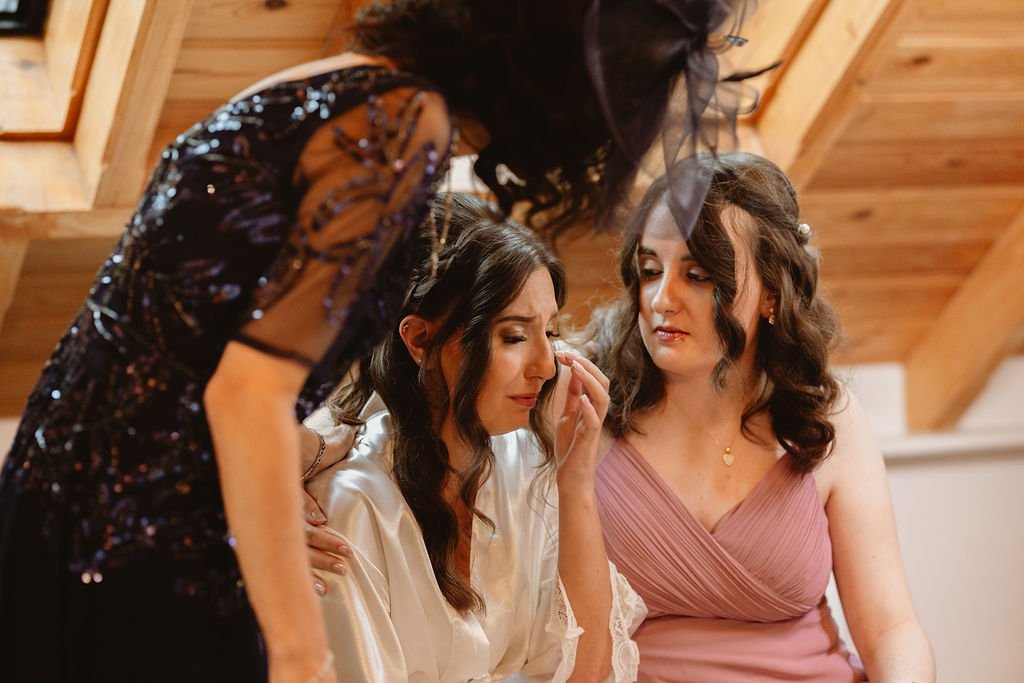 A woman with long dark hair and a white satin robe is crying while another woman with shoulder-length wavy brown hair in a pink dress gently wipes her tears, as a third woman with long dark hair in a black sequined dress looks on, inside a wooden-pan