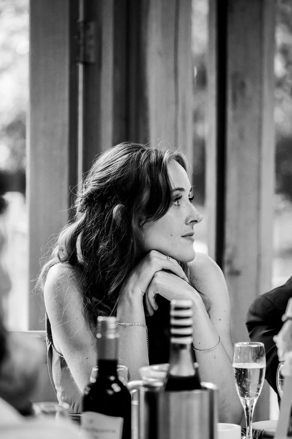 A young woman with wavy hair, sitting at a dining table, resting her chin on her hands, gazing to the right. The table has a wine bottle and a glass of champagne in front of her.