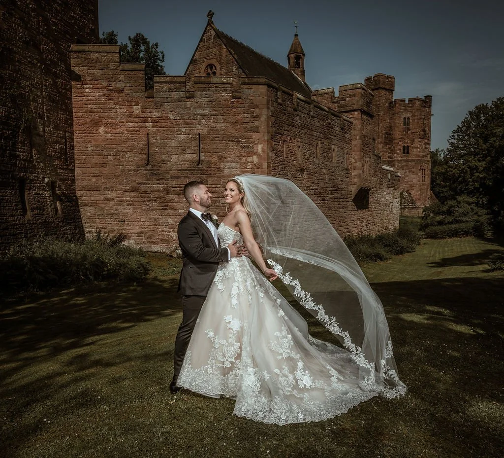 Bride and groom in wedding attire standing close together on a grassy area outdoors, with a historic stone castle in the background during daytime.