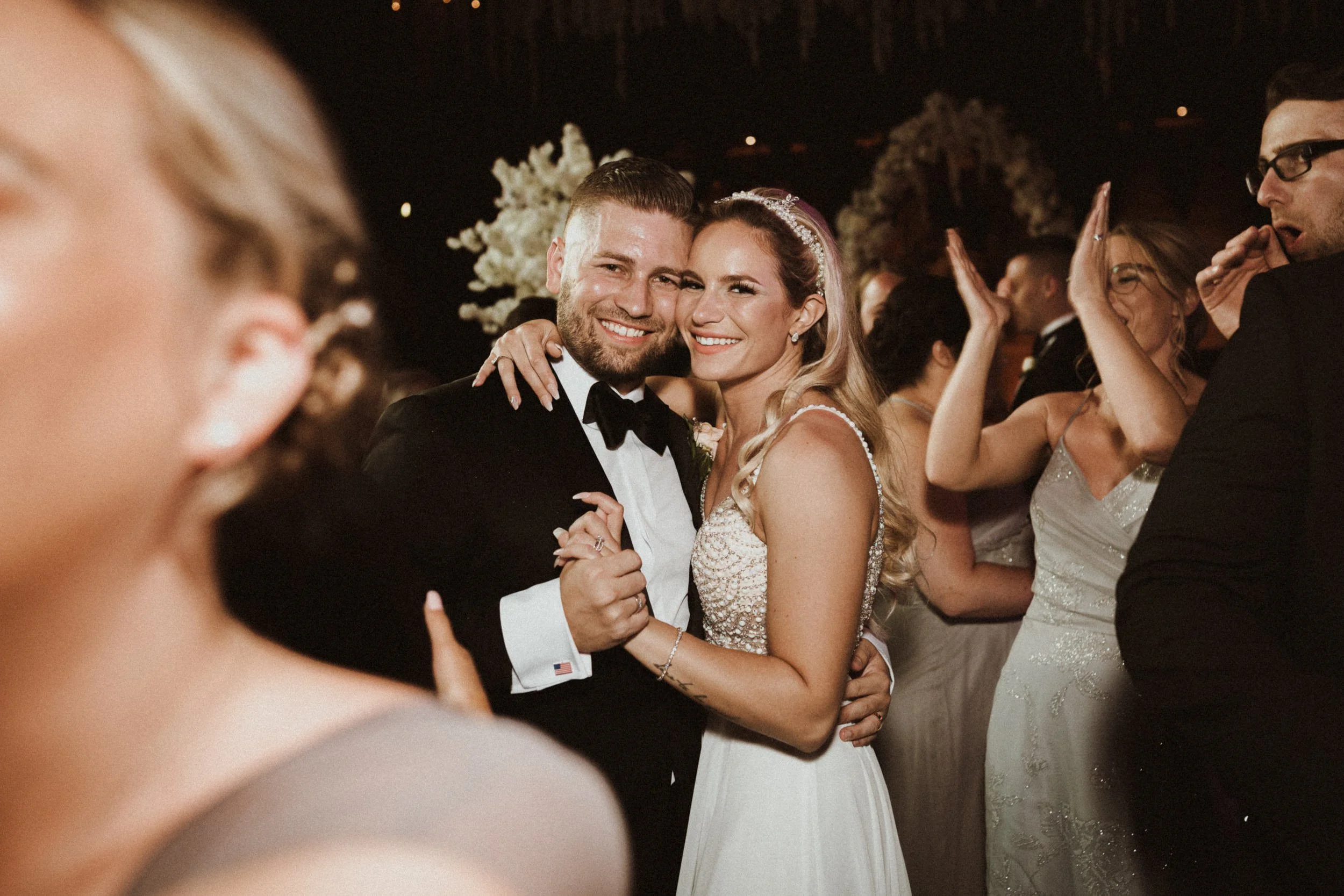 A newlywed couple in wedding attire smiling and holding each other on the dance floor at their wedding reception, surrounded by friends and family.