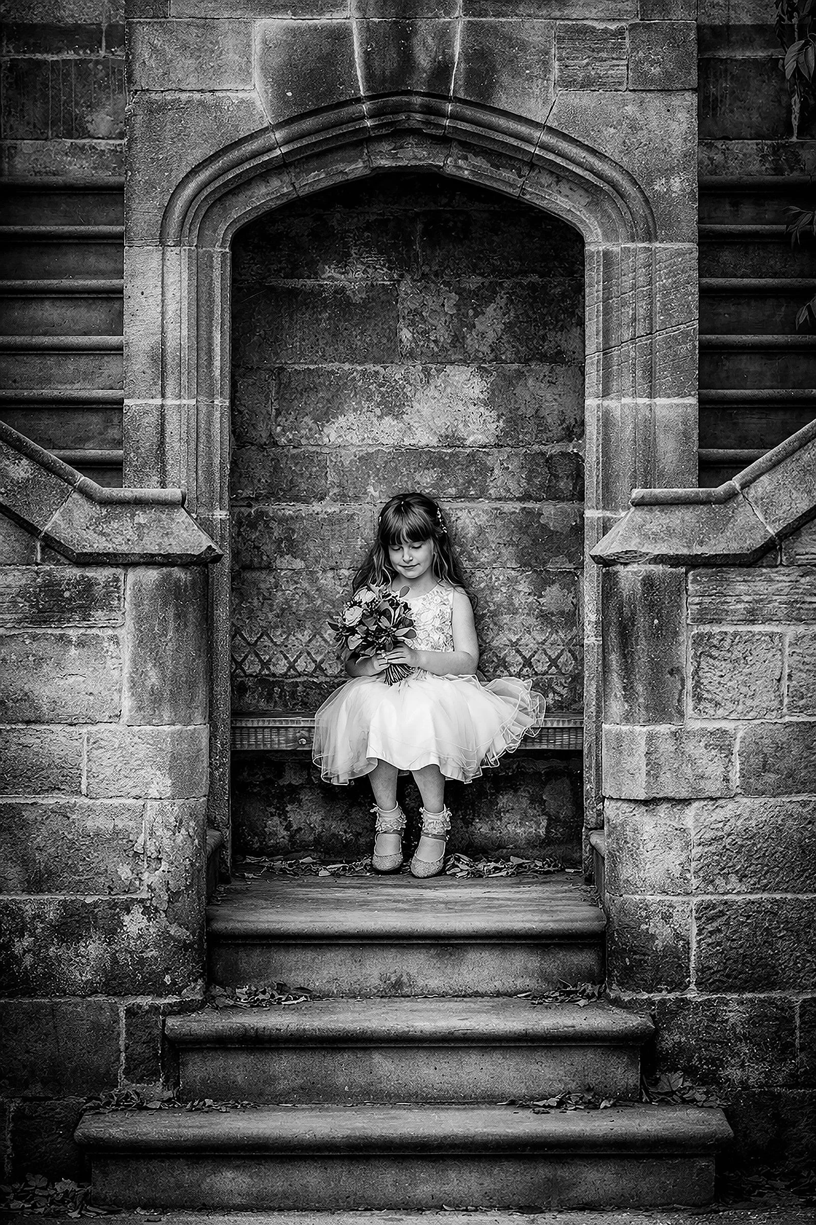 Black and white photo of a young girl in a dress holding a bouquet of flowers, sitting on a bench within an old stone archway at the top of stone stairs.