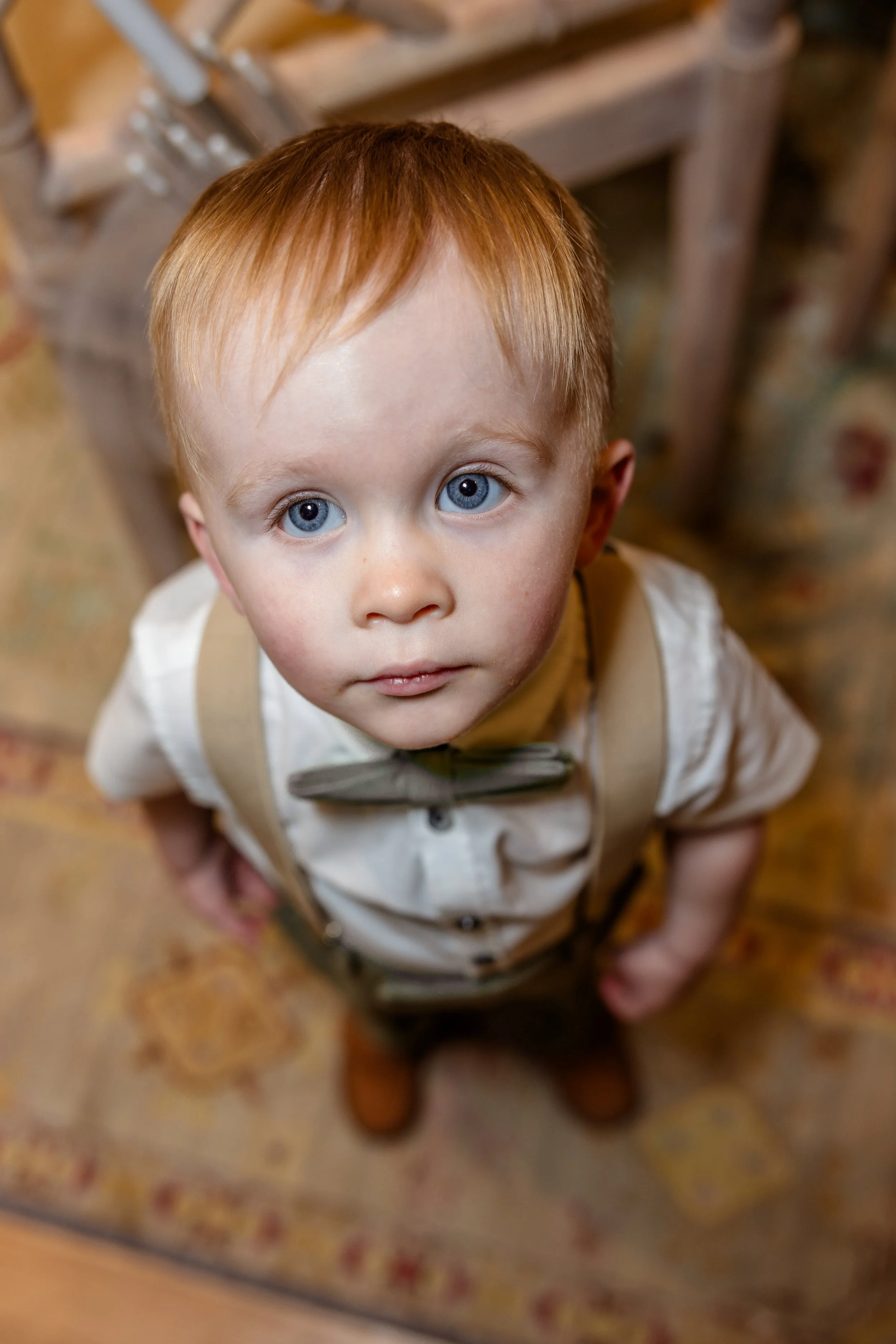 A young boy with red hair and blue eyes looking up at the camera, dressed in vintage-style clothing including a white shirt, suspenders, a bow tie, and brown shoes, standing on a patterned rug.