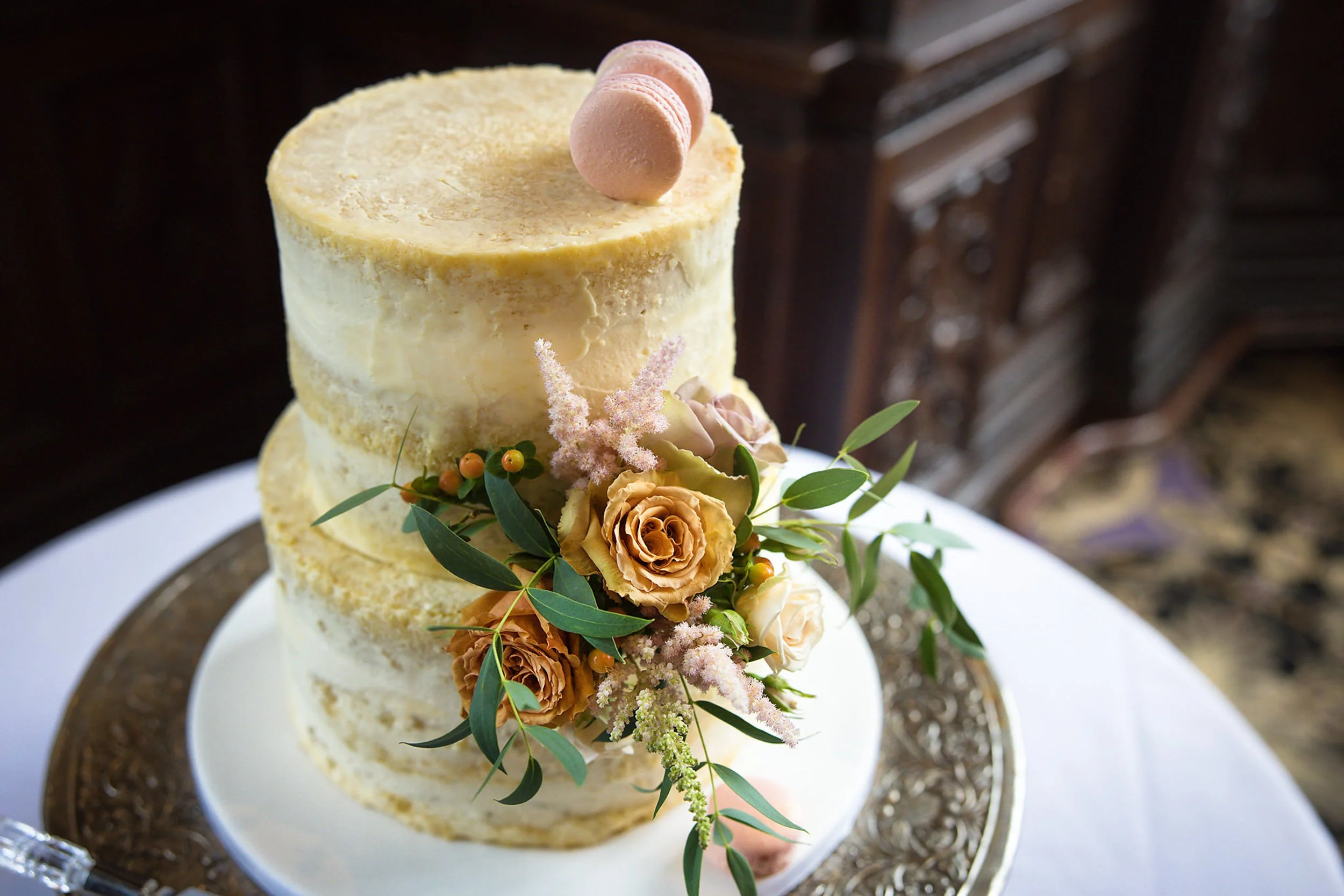 A three-tier semi-naked wedding cake decorated with a flower arrangement including roses and greenery, topped with pink macarons, placed on a silver-edged cake stand.