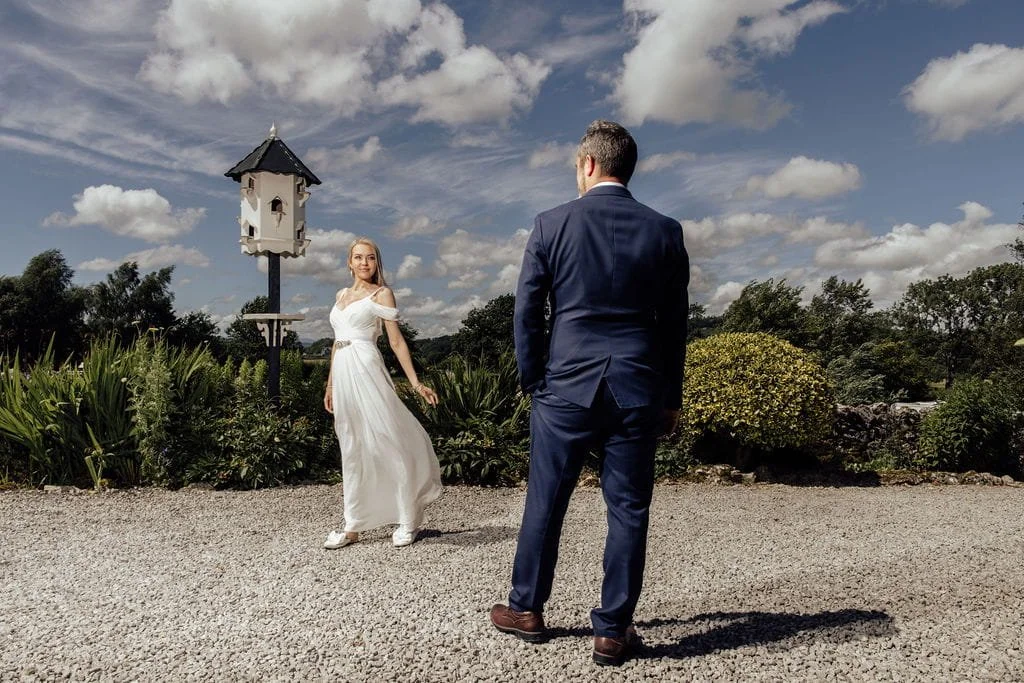 A man in a blue suit faces a woman in a white dress outdoors, with a garden, bushes, and a birdhouse on a pole in the background under a partly cloudy sky.