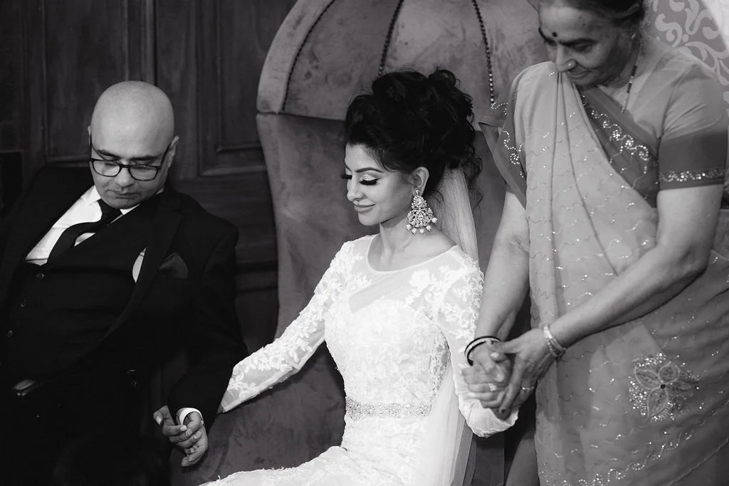 A bride dressed in a white lace wedding gown, sitting with her eyes closed and smiling, holding hands with two people, one man in a suit and woman in a traditional sari, during a wedding ceremony.