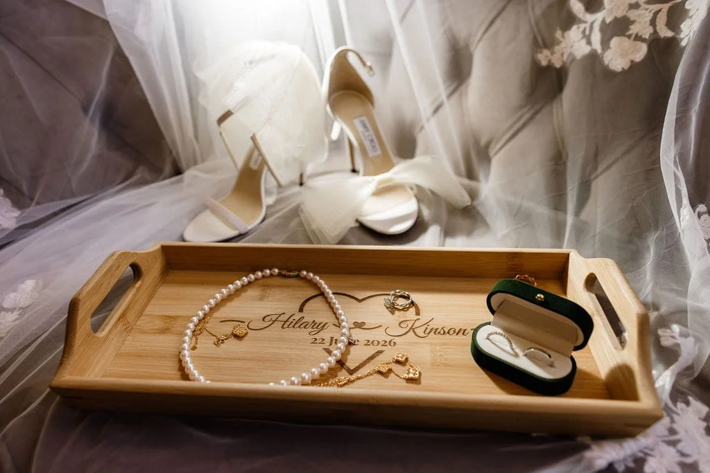 Wedding jewelry and accessories on a wooden tray with the names Hilary and Kayson and the date October 22, 2026, written on it, with white wedding shoes in the background.