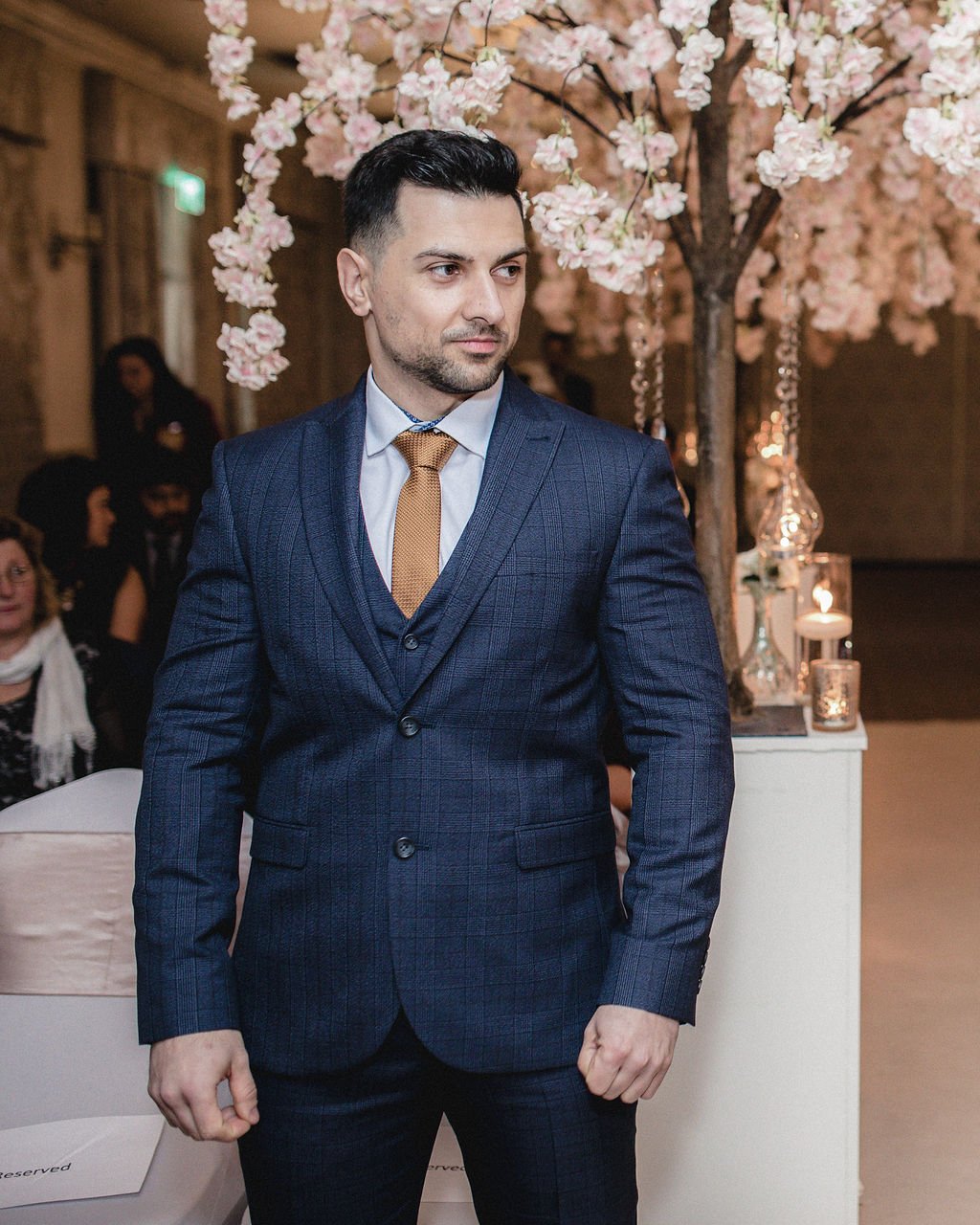 A man in a dark blue checked suit with a light blue shirt and a gold tie standing in front of pink cherry blossom trees in an indoor event setting, with candles and guests in the background.