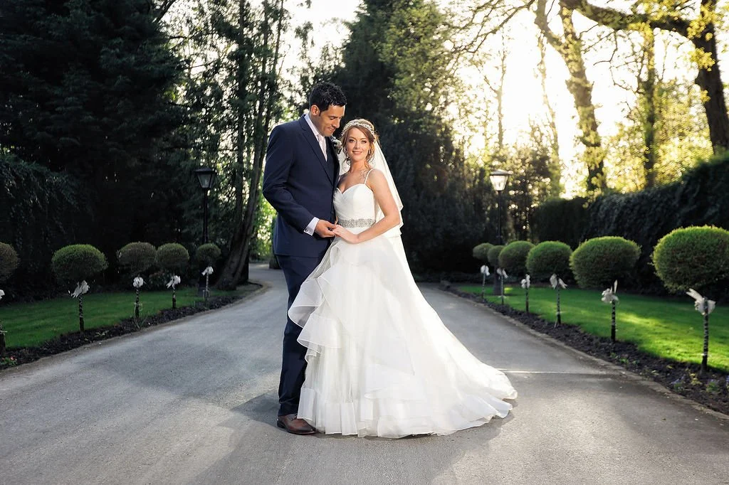 A bride and groom standing close together on a paved pathway in a park, surrounded by green trees and bushes, with sunlight filtering through the leaves.