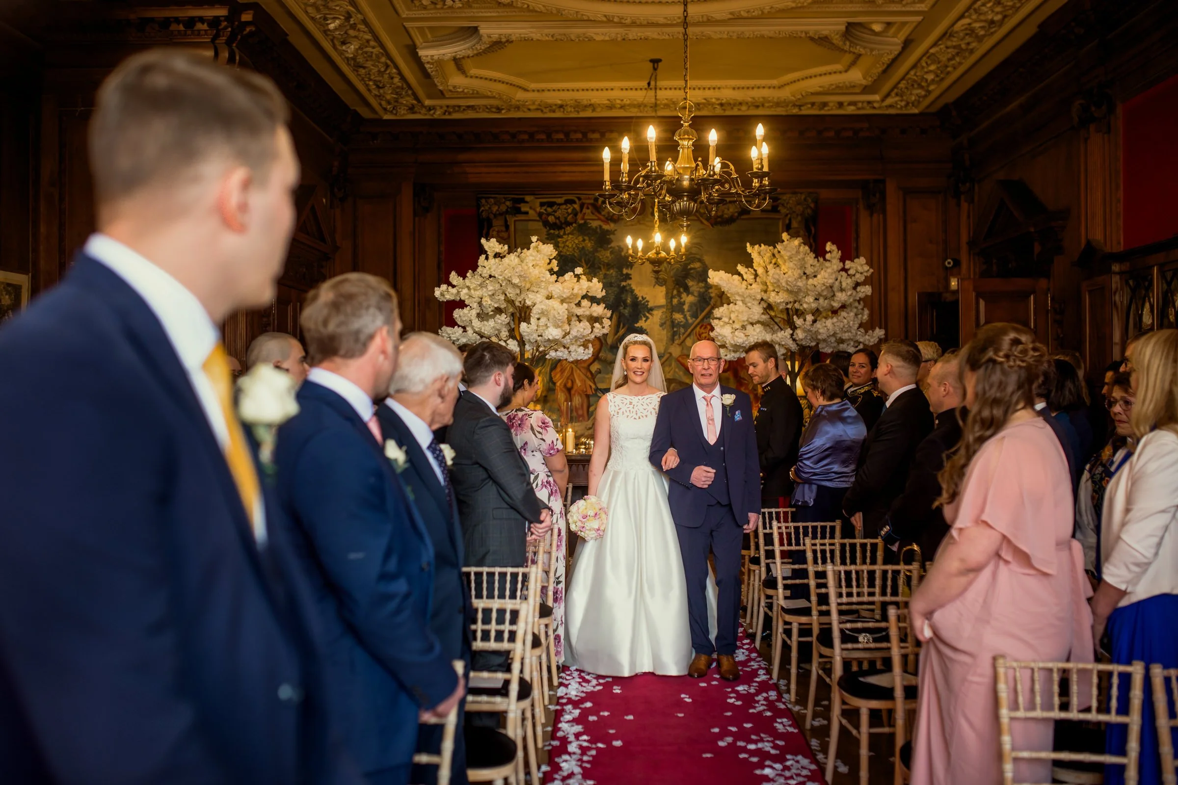 A bride walking down the aisle escorted by an older man at her wedding ceremony. Guests are seated on both sides of the aisle, and decorated with white flowers and petals. The room has ornate wood paneling, a chandelier, and large cherry blossom tree