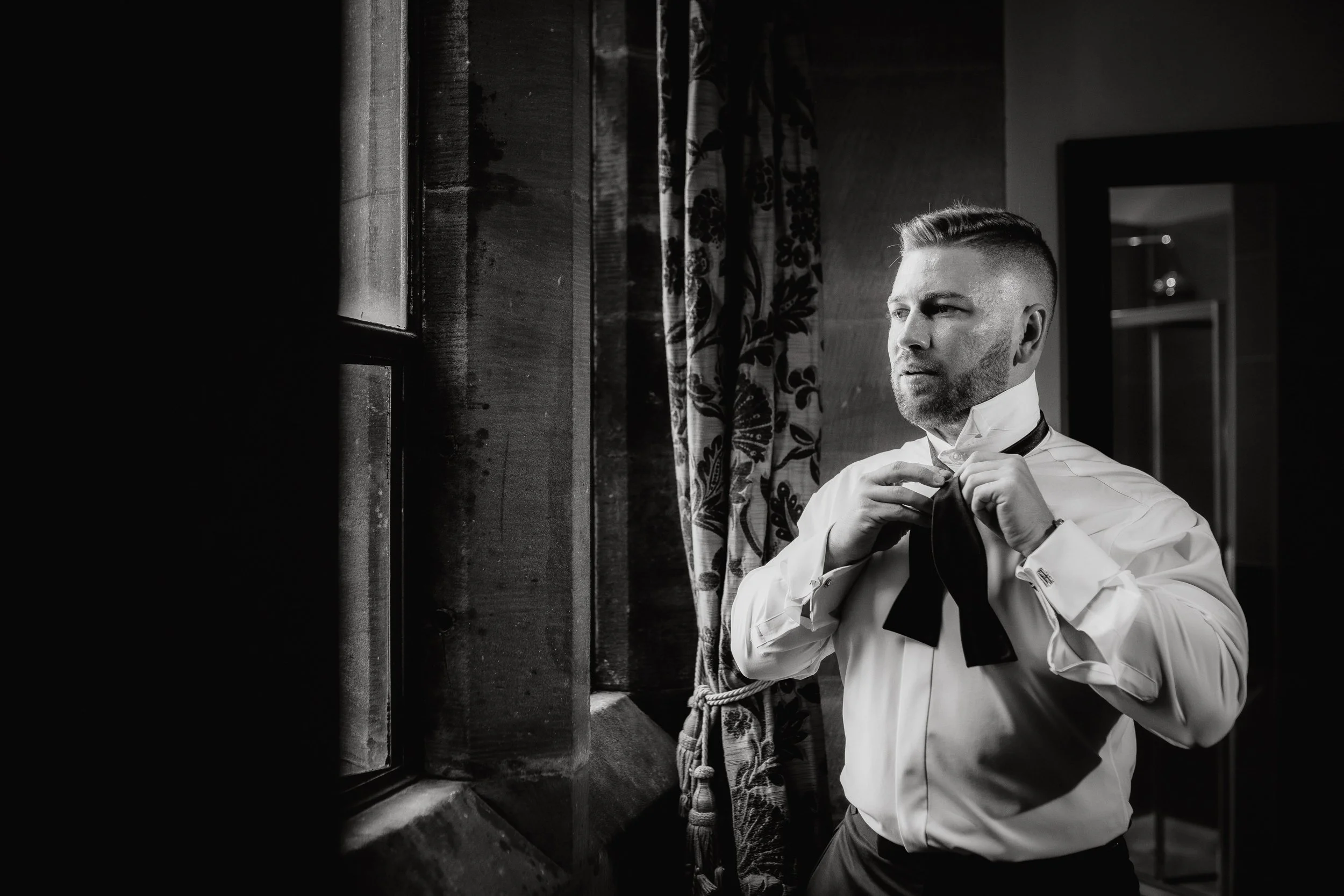 A man adjusts a necktie in front of a window, with curtains behind him, in a room with dark wooden interior.