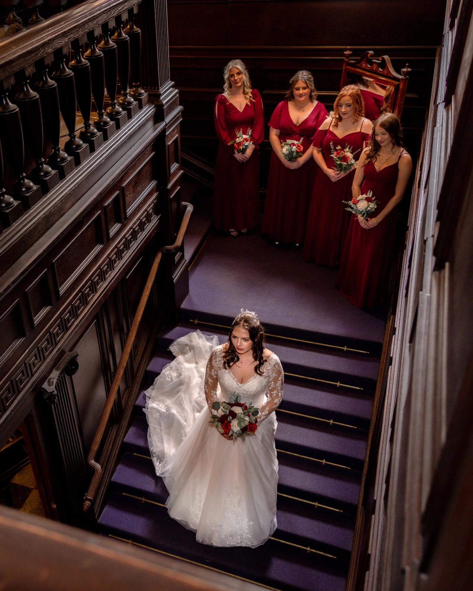 Bride sitting on stairs with bouquet, surrounded by four bridesmaids in red dresses holding bouquets, in an elegant wooden staircase setting.