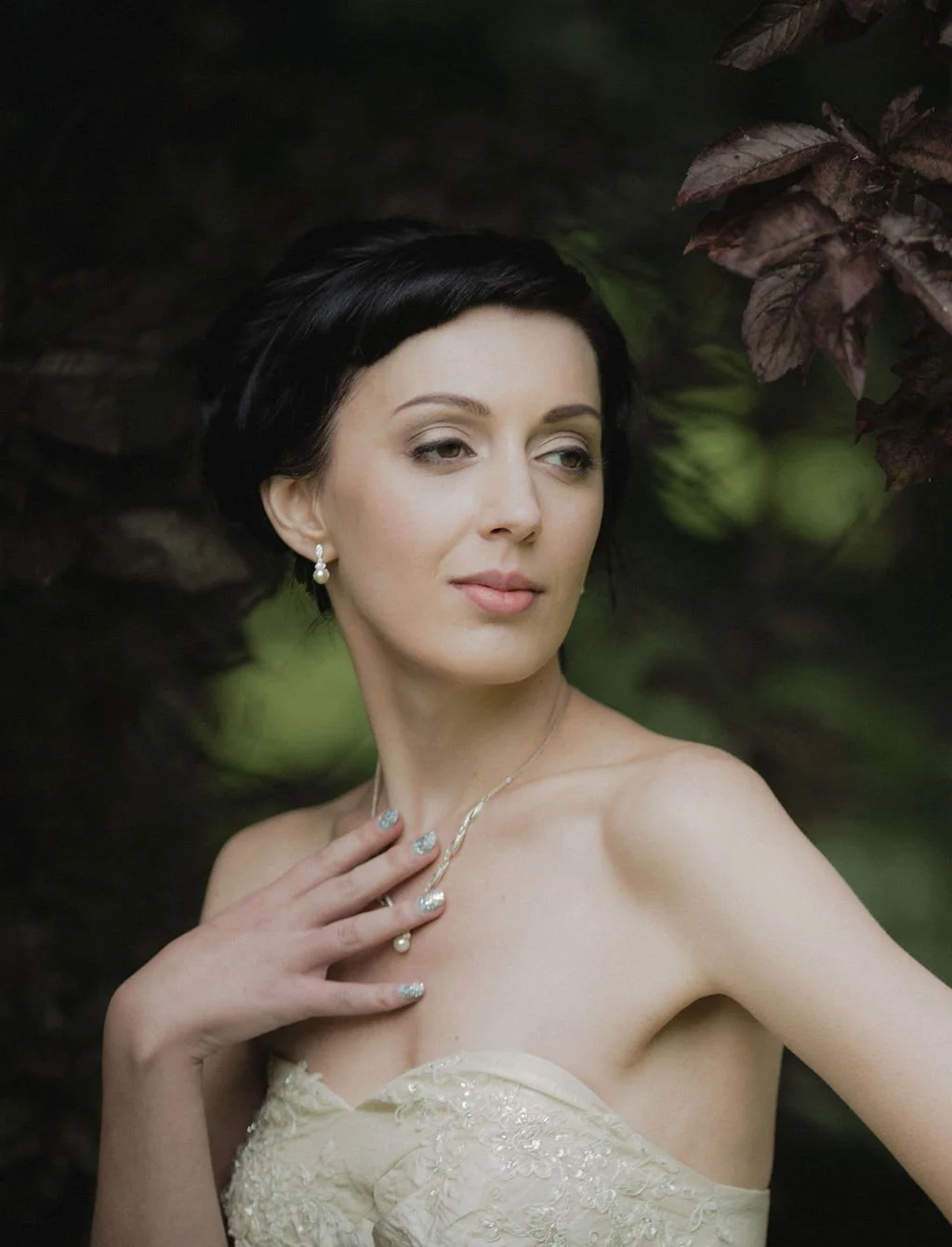 A woman with short black hair and pearl earrings poses outdoors, wearing a strapless beige dress with lace details, showing her shoulder and necklace, with a background of dark leaves and greenery.