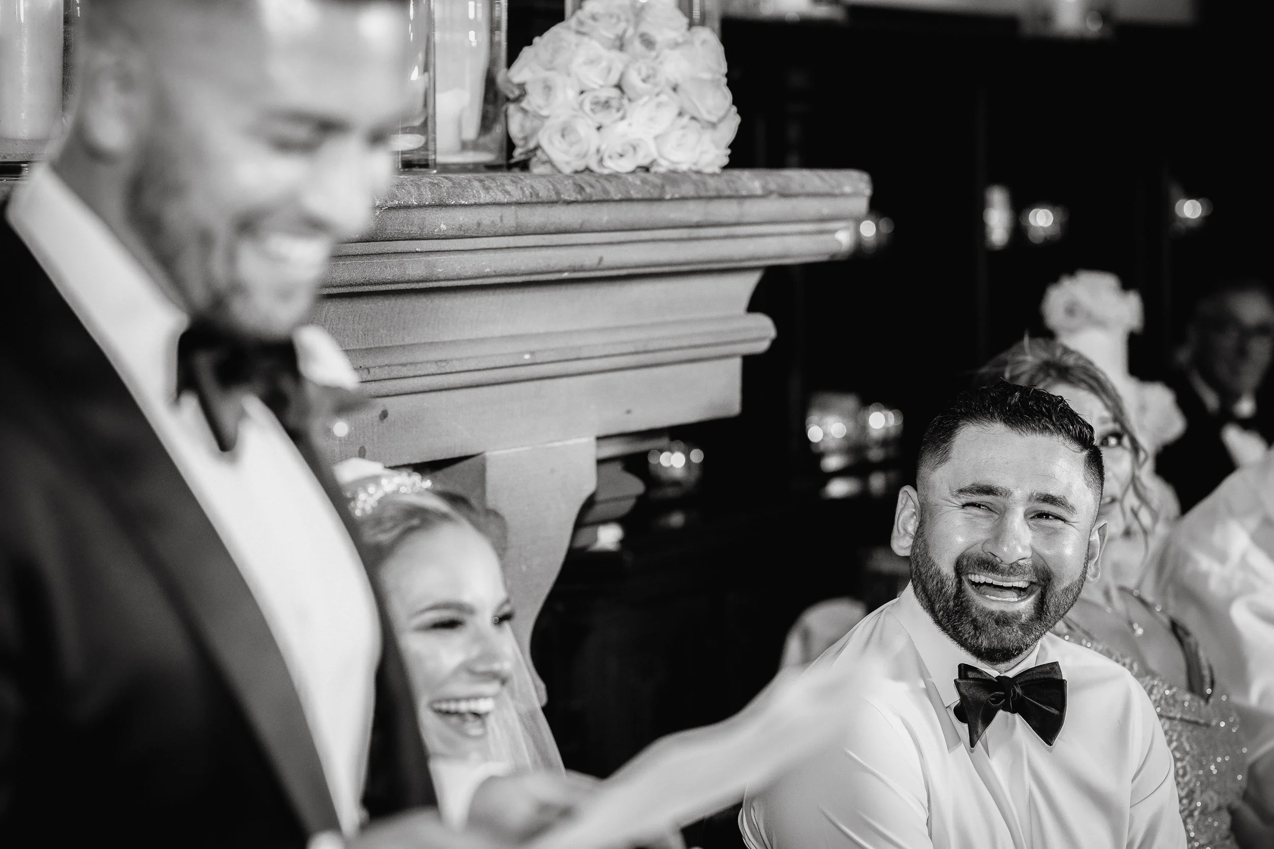 A black and white photo of people dressed in formal attire, including tuxedos and dresses, sitting together and smiling, likely at a wedding or celebration.